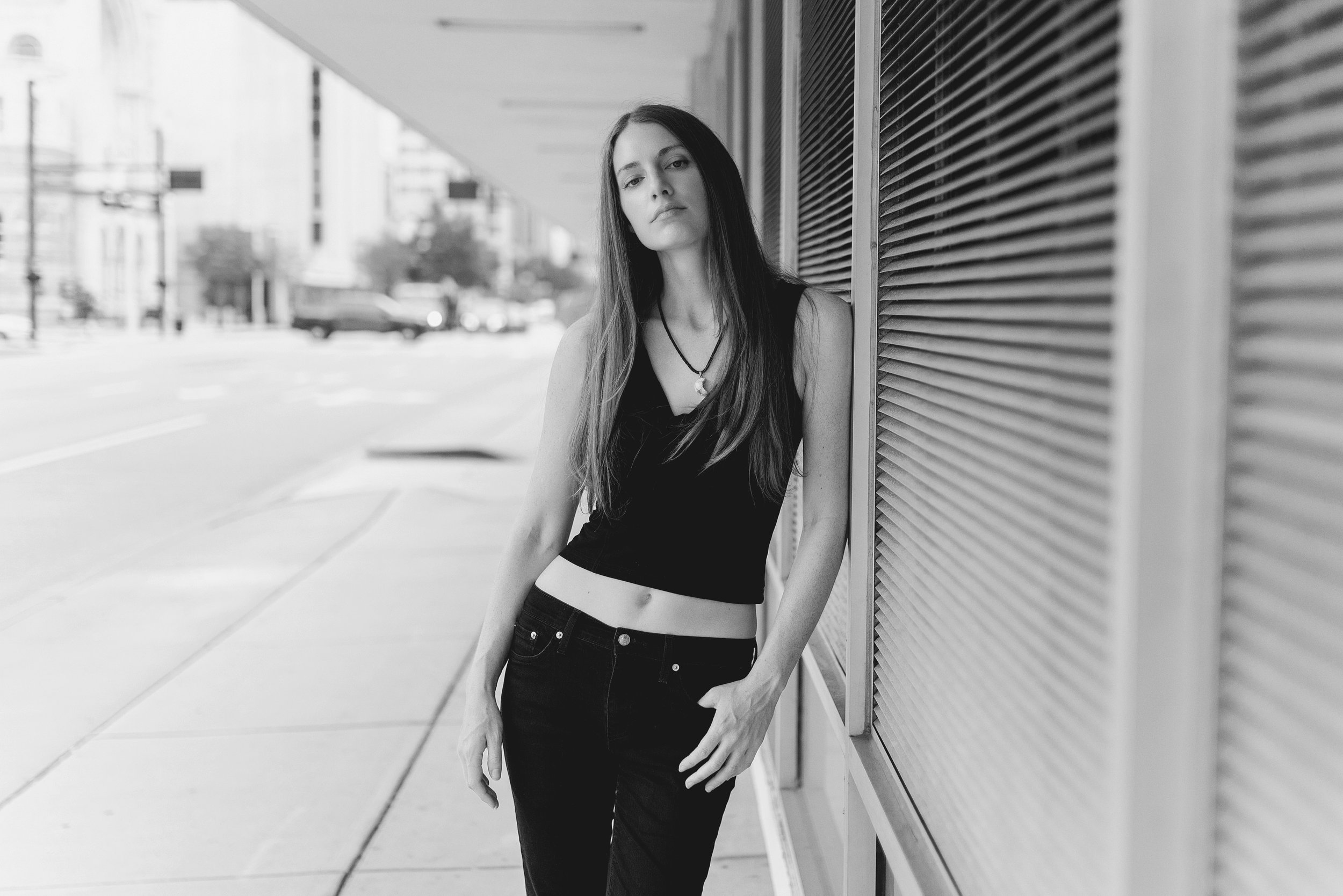 A young woman with long dark hair, wearing a black sleeveless top and black jeans, stands against a wall with horizontal slats on an urban sidewalk. She has a serious expression and looks directly at the camera, in Tampa, FL.