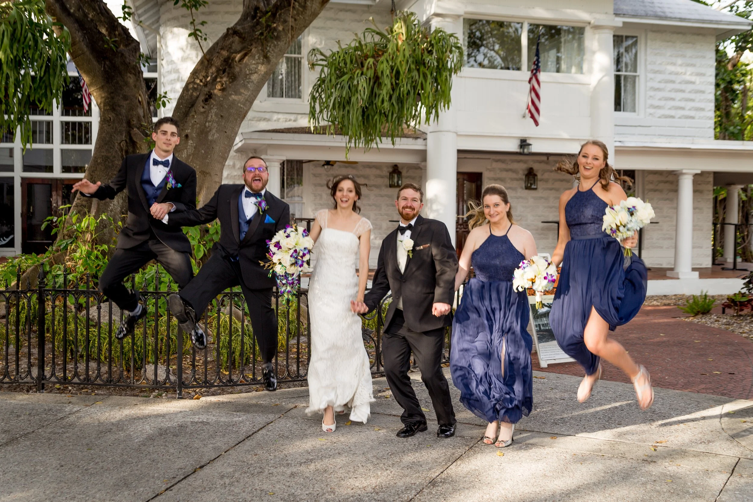 Group of six people, including a bride and groom, celebrating outside a white house with American flags. Two women in matching blue dresses and two men in black suits with bow ties are smiling and jumping, holding bouquets of flowers.