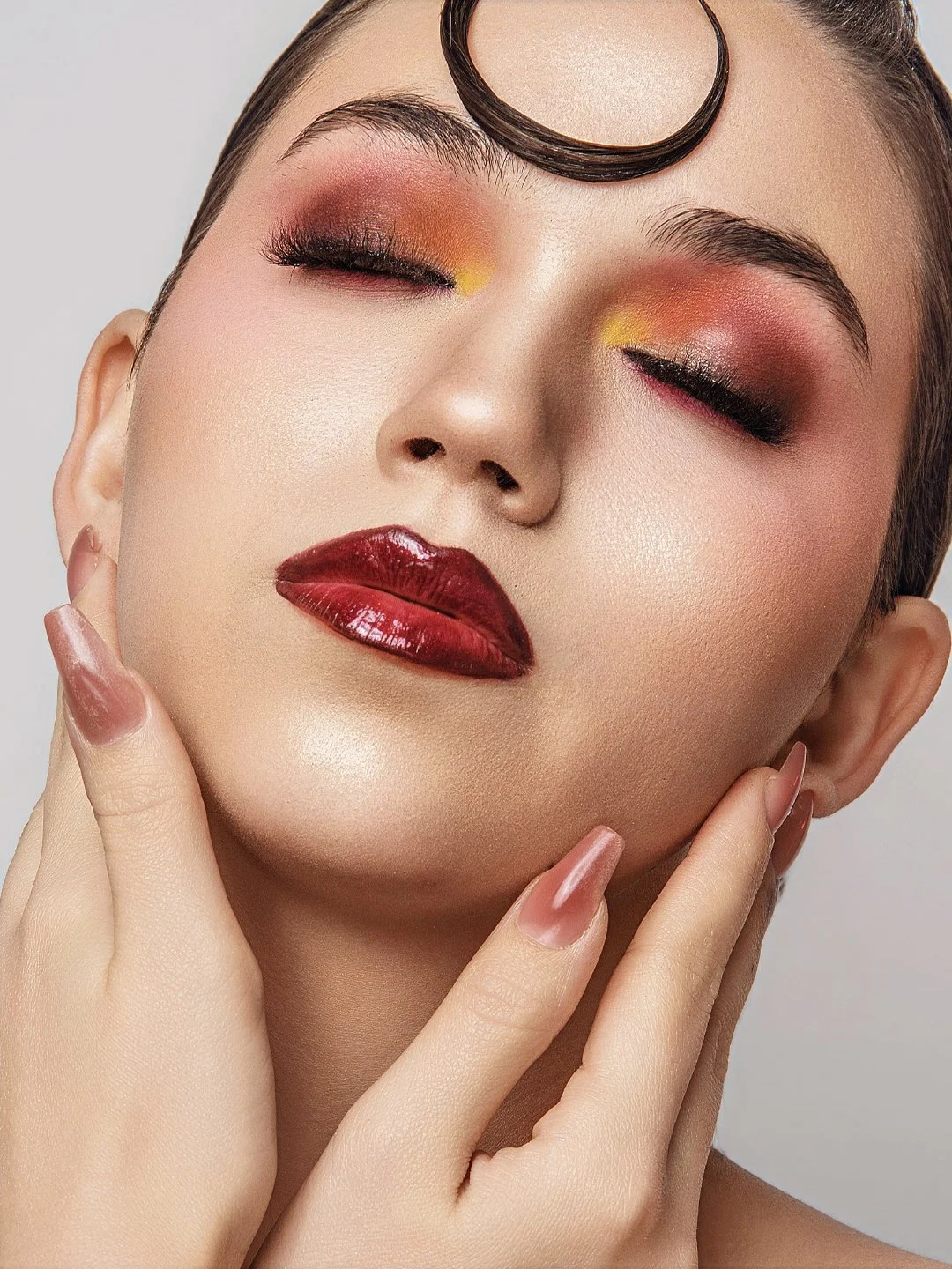 Close-up of a woman with bold makeup, including glossy dark red lipstick, shimmery eye shadow in shades of red, yellow, and black, and well-groomed eyebrows. She is gently touching her face with her hands, and has a curl of hair on her forehead.