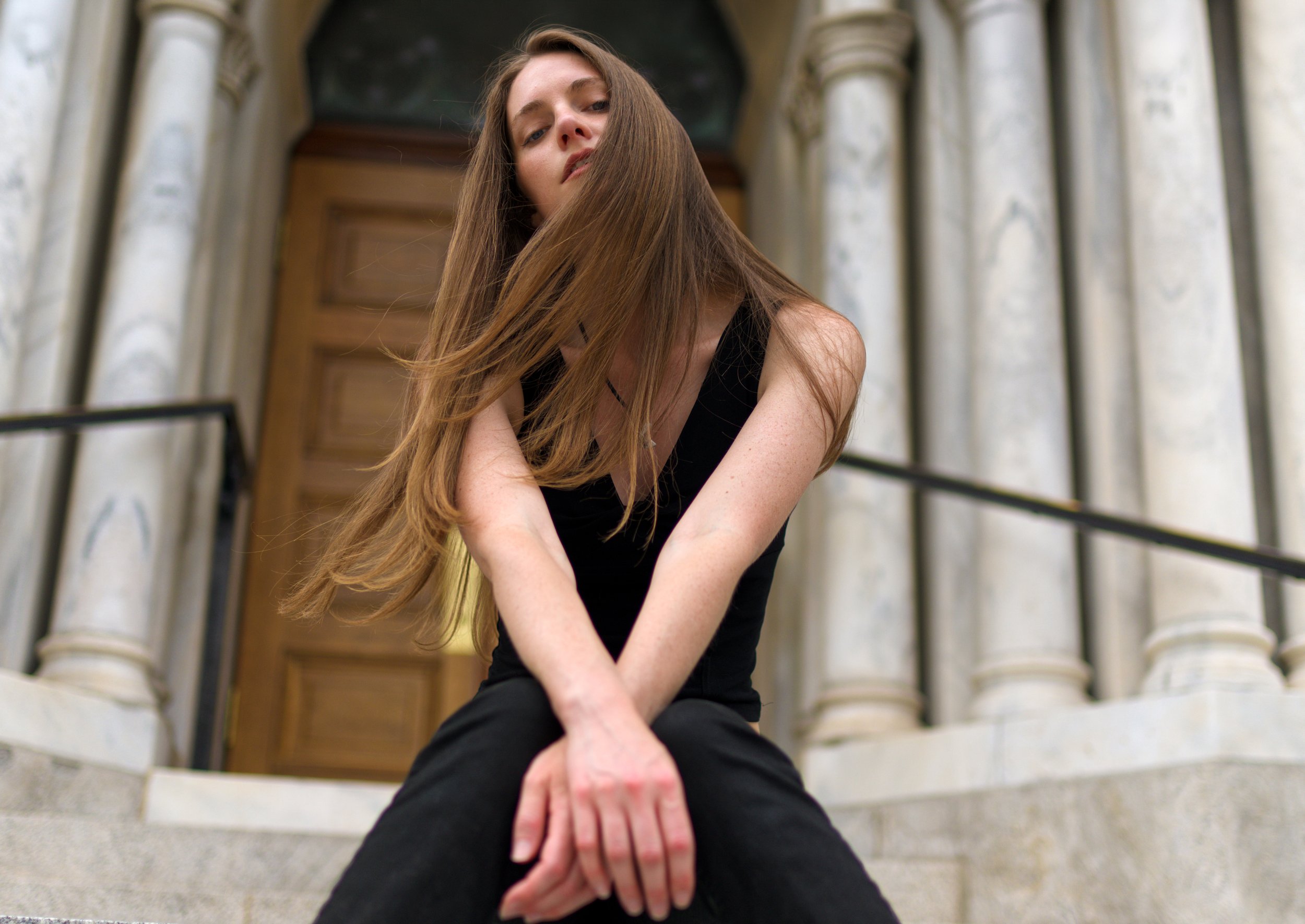Young woman with long brown hair sitting on stone stairs in front of a building with columns, looking at the ,  in Tampa, FL