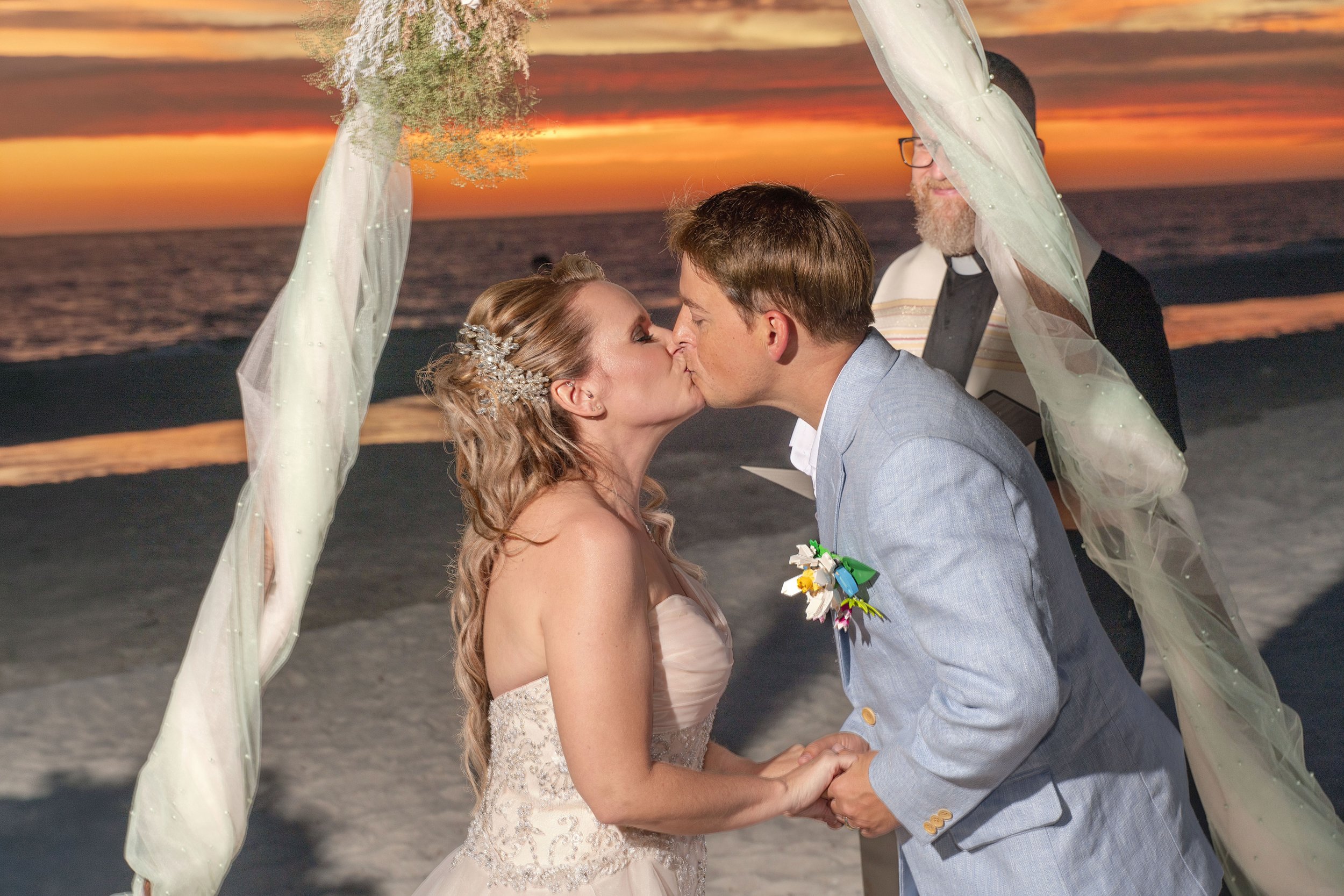 A bride and groom kiss at their wedding ceremony on the beach during sunset, with a preacher officiating in the background.