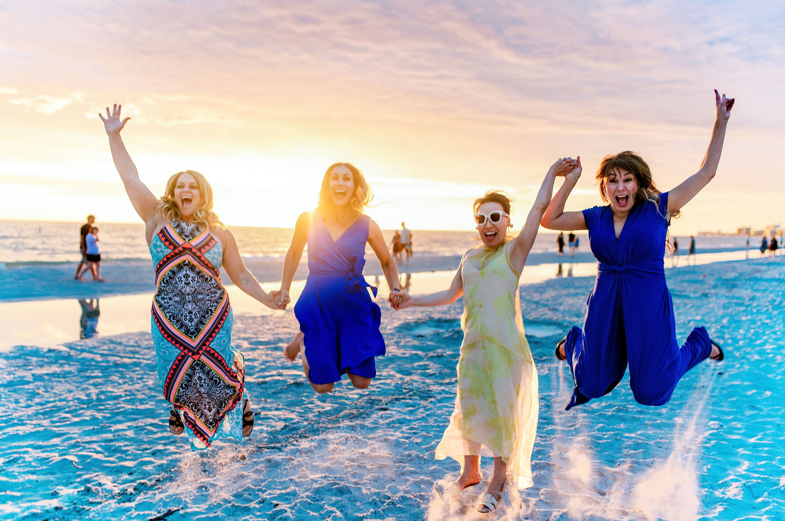 Four women holding hands and jumping on the beach during sunset, wearing colorful dresses and sunglasses, with the ocean and other beachgoers in the background.
