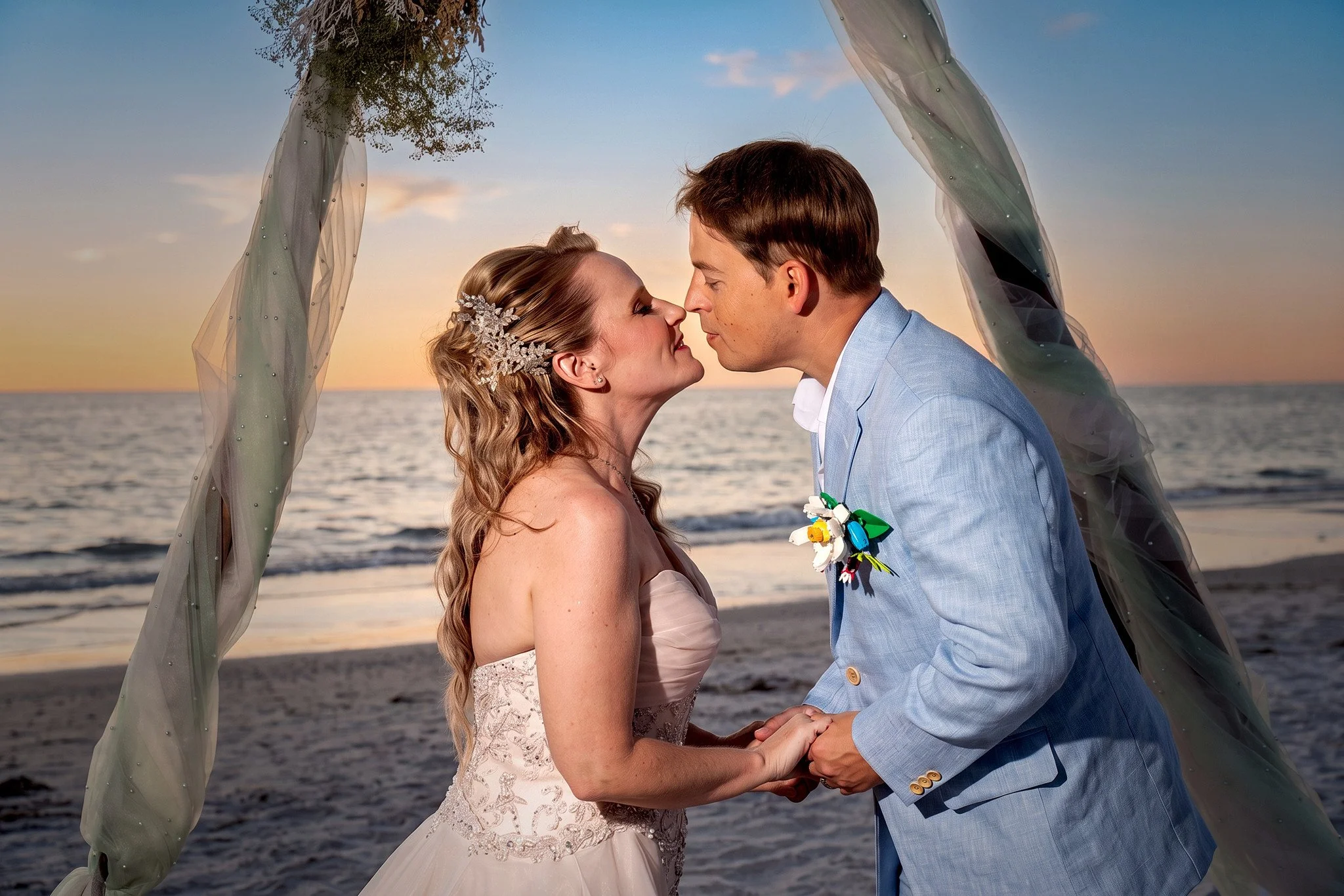 A bride and groom holding hands and about to kiss on a beach during sunset, standing beneath a decorative arch with fabric and greenery.