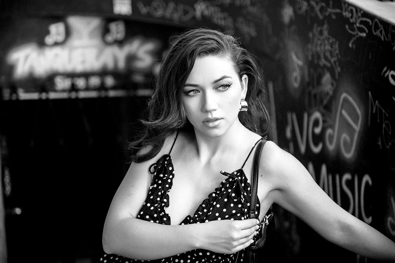 A black-and-white photo of a young woman with wavy hair, wearing a polka dot dress with thin straps, standing next to a chalkboard wall with music and tattoo-related writings.