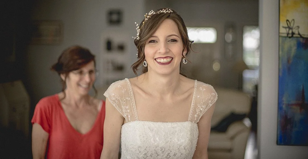 A bride smiling, wearing a white wedding dress with lace detailing and a pearl headband, standing indoors with an older woman in a red top behind her.