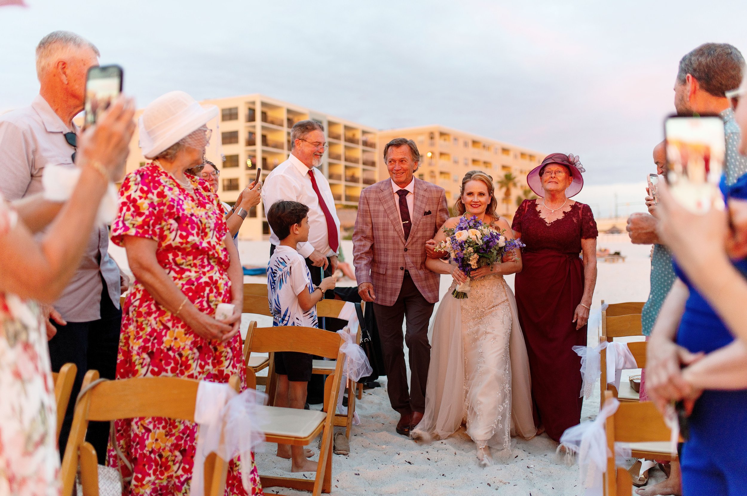 A bride holding a bouquet walking down the aisle at a beach wedding, surrounded by seated guests and wedding attendees, with modern apartment buildings in the background during sunset.