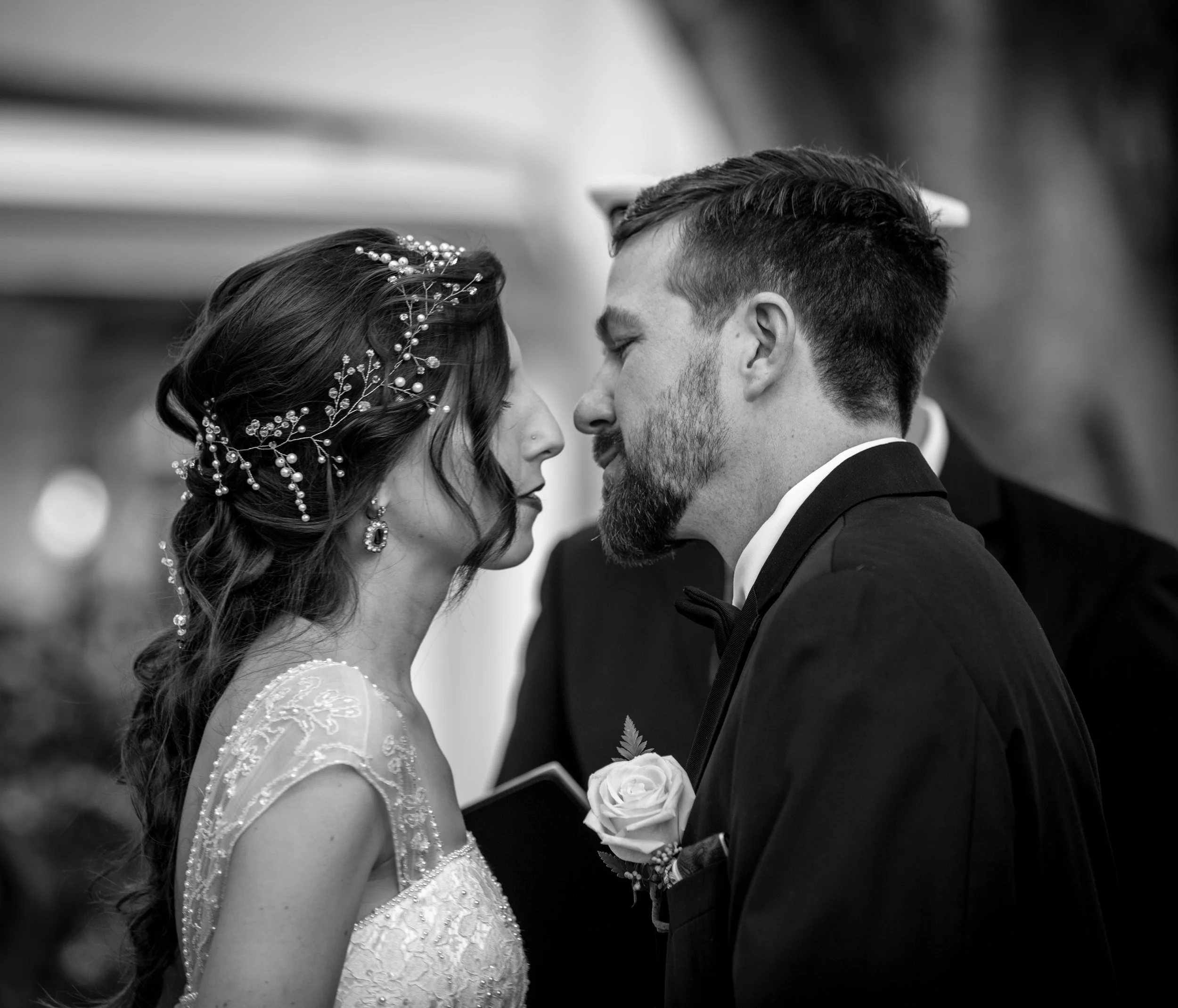 A black-and-white photo of a bride and groom close together, touching foreheads, with their eyes closed. The bride wears a lace wedding dress with embroidery and a hairpiece with pearls, while the groom wears a tuxedo with a white rose boutonniere.