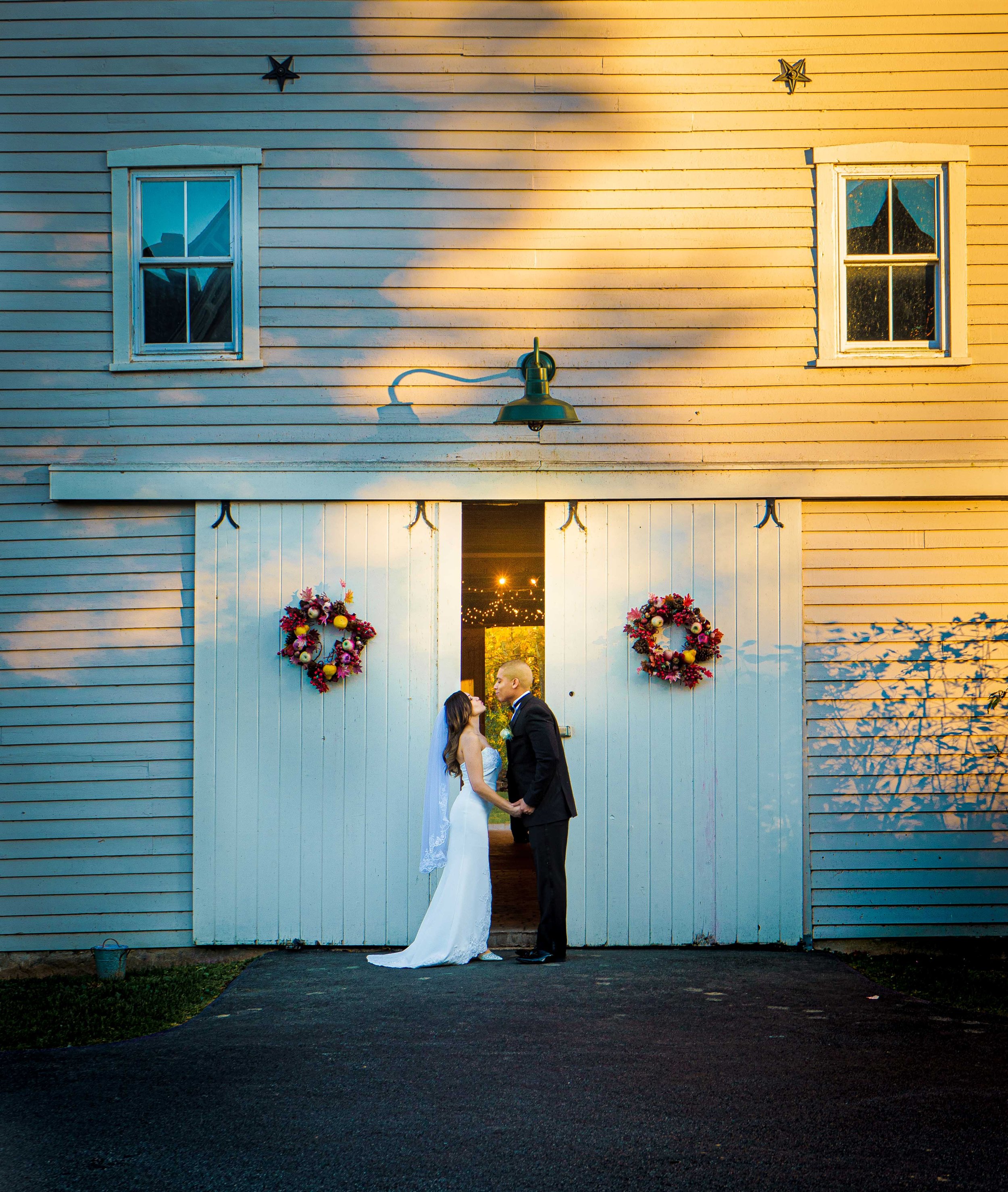A bride and groom holding hands and facing each other in front of a large white barn with two windows and decorated with two floral wreaths. There are star-shaped decorations on the barn. The scene is lit with warm sunlight and there are lights visib
