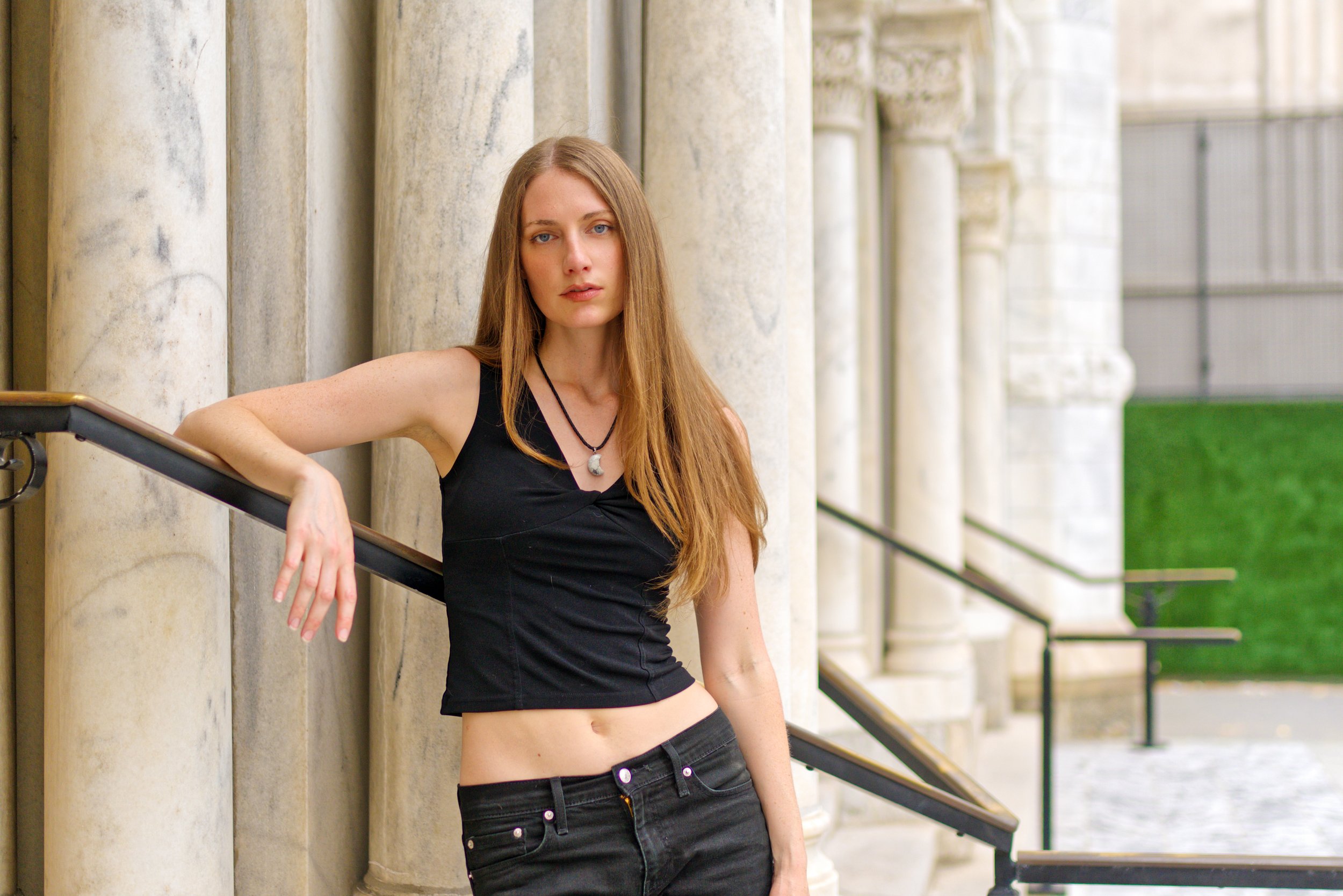 A young woman with long red hair and blue eyes, wearing a black tank top and black pants, leaning against marble columns outdoors,  in Tampa, FL.
