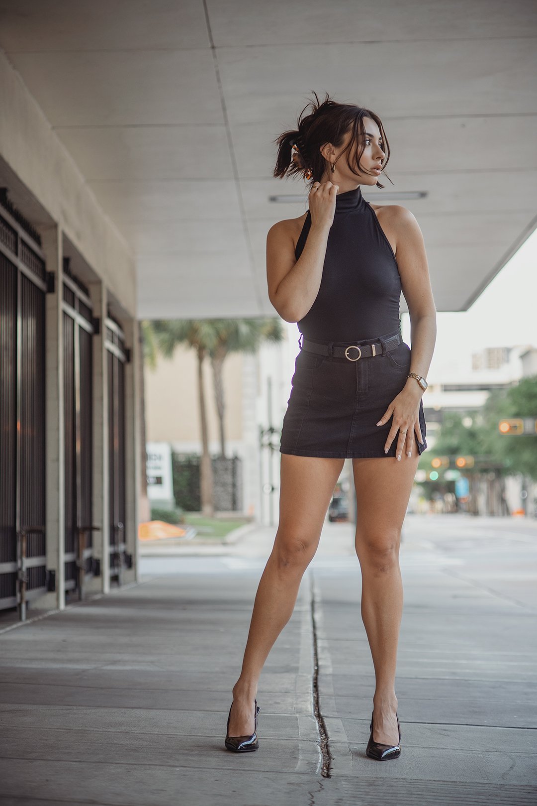 A woman with short dark hair wearing a sleeveless black turtleneck top, a black mini skirt with a belt, and black high heels, standing on a city sidewalk with trees and buildings in the background.