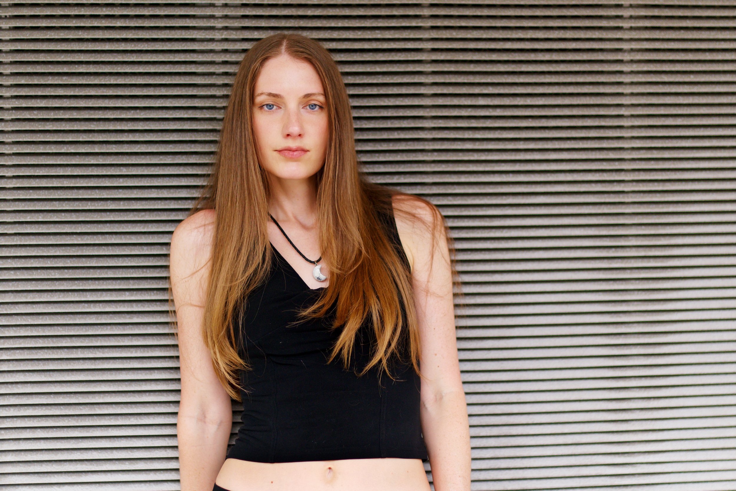 A young woman with long red hair and blue eyes standing against a horizontally striped gray wall, wearing a black sleeveless top and a moon-shaped necklace,  in Tampa, FL.