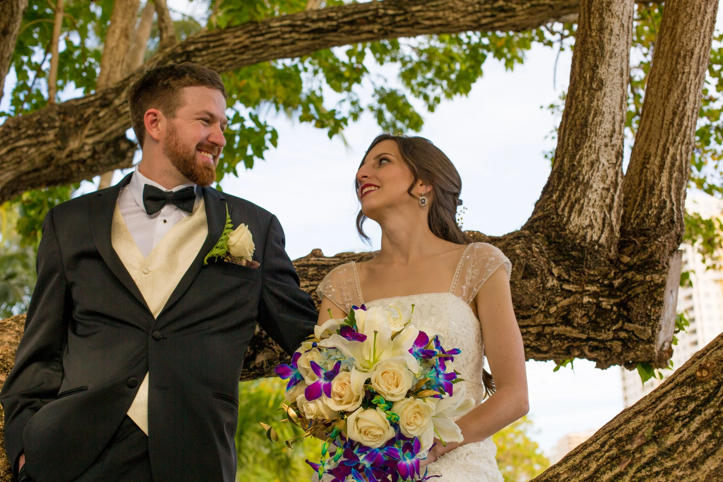 A bride and groom looking at each other, standing under a large tree outdoors, with the bride holding a bouquet of white and purple flowers.