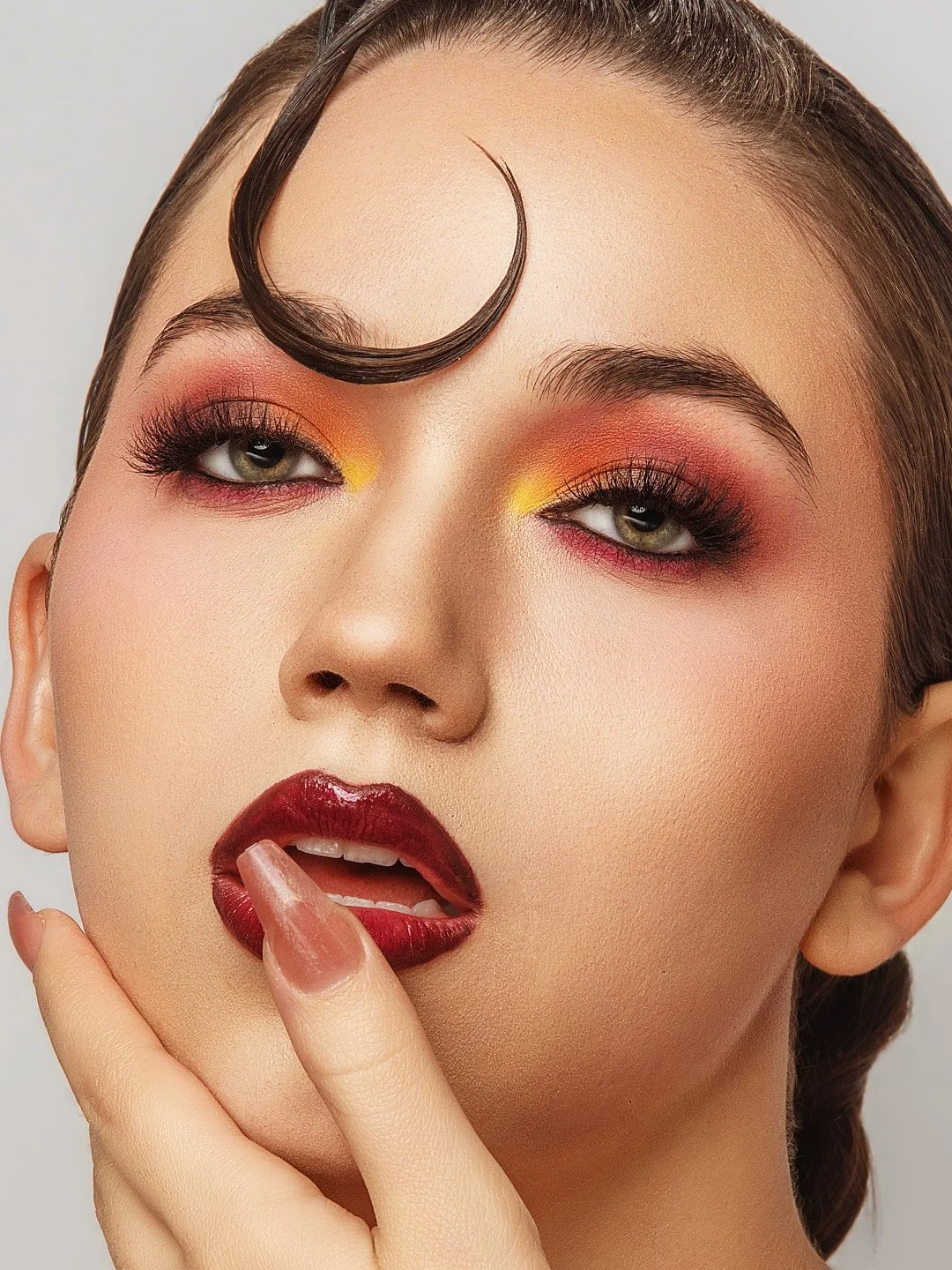 Close-up of a woman with bold makeup, dark red lipstick, shiny nails, eye makeup with red and yellow eyeshadow, and a curled strand of hair on her forehead.