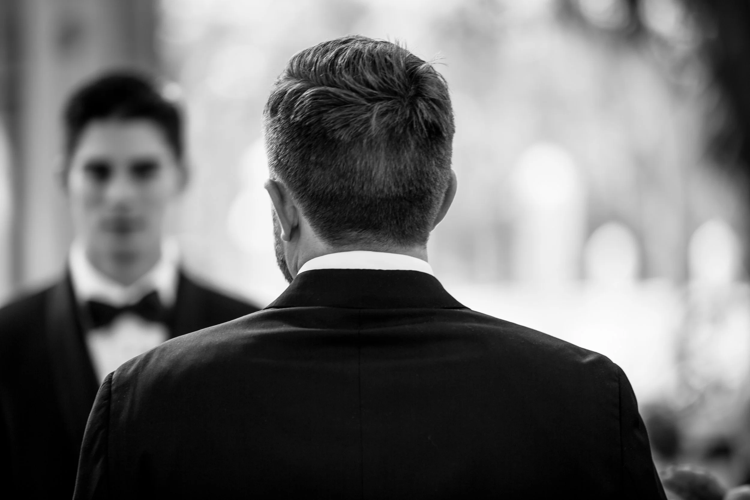 Man with short hair from behind, wearing a suit, facing a woman with short hair and glasses in a blurred background.