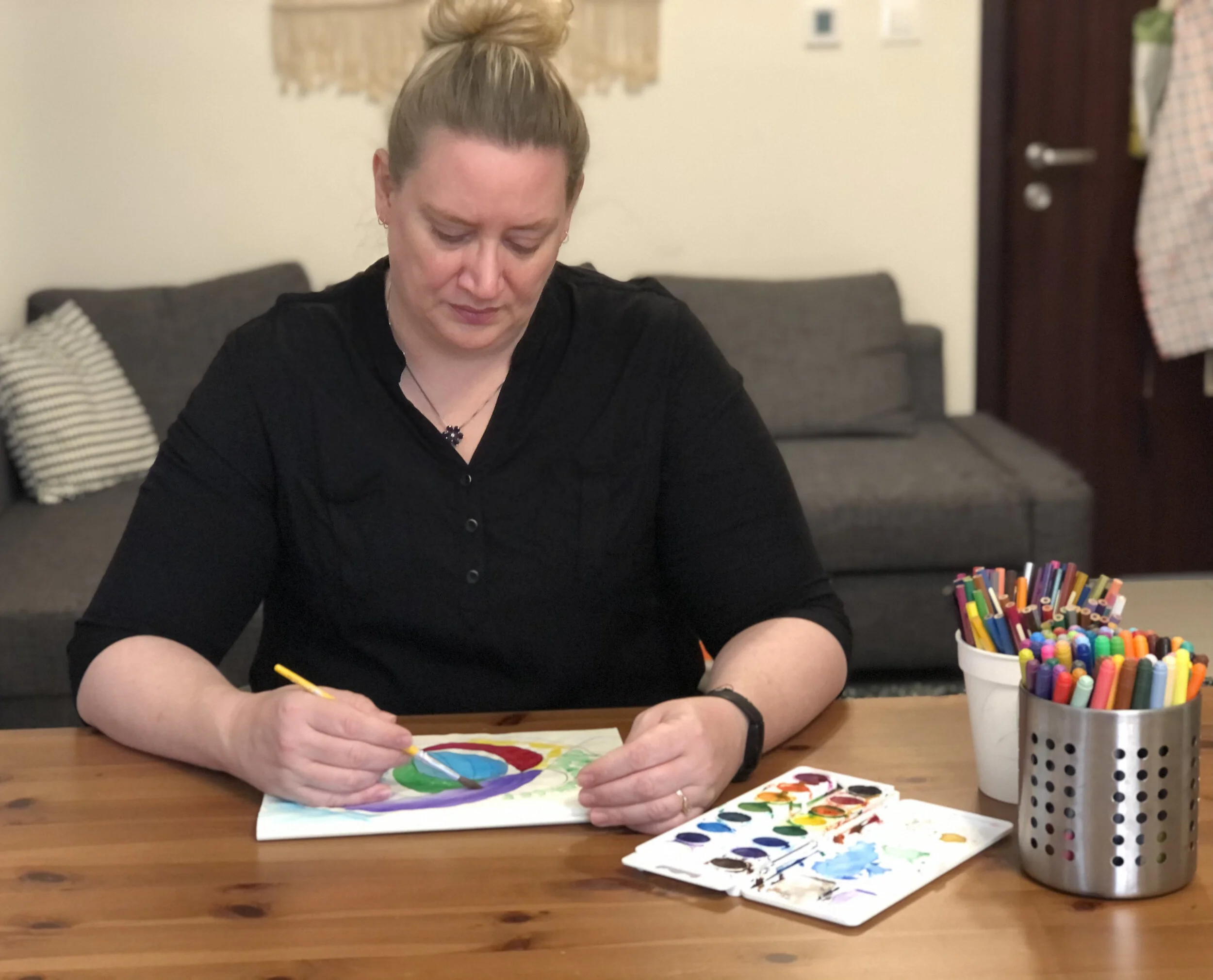 A woman with blonde hair tied in a bun, wearing a black shirt, paints a colorful rainbow on paper using a small brush. There is a palette with various watercolor paints in front of her, and two containers filled with colorful markers on the table. Th