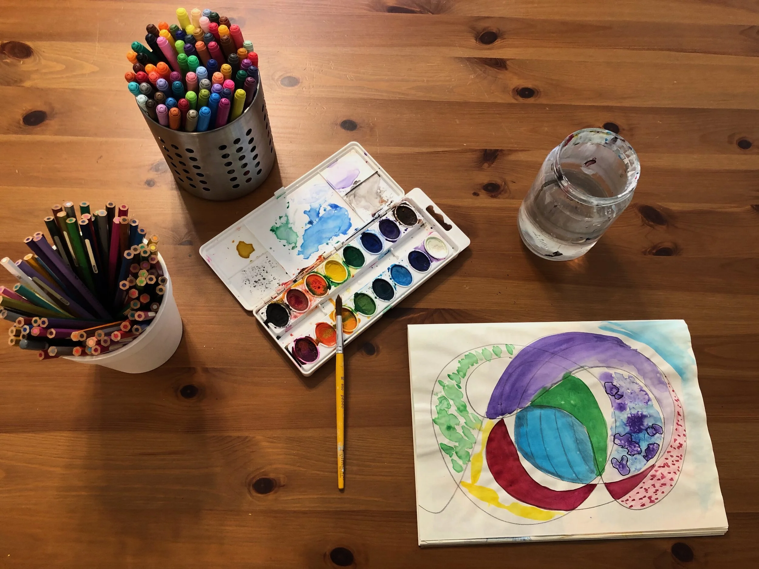 A wooden table with containers of colored pencils, watercolor paints and a brush, a glass of water, and a watercolor painting of abstract colorful shapes.
