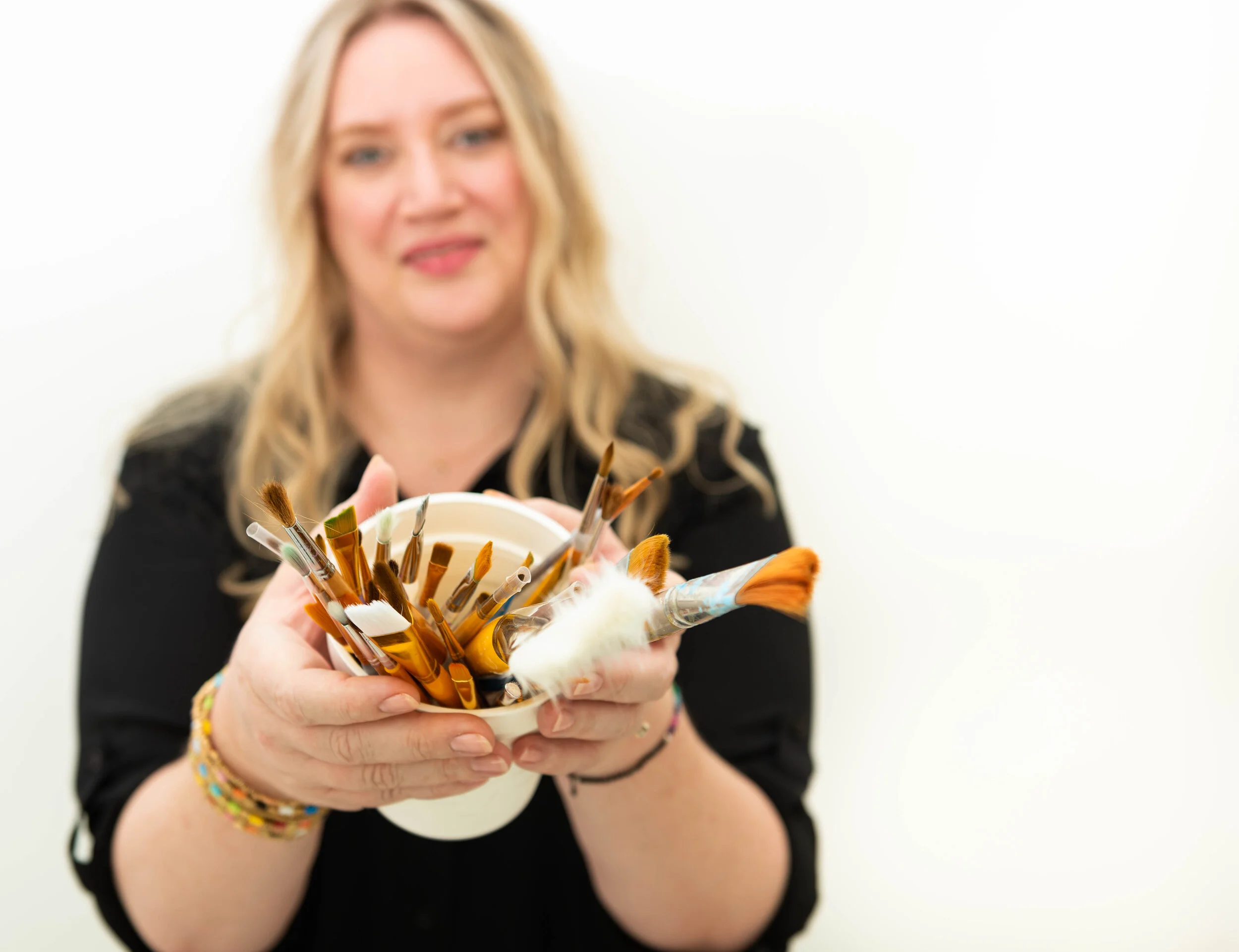 A woman with blonde hair wearing a black shirt is holding a white cup filled with paintbrushes, presenting it towards the camera against a plain white background.