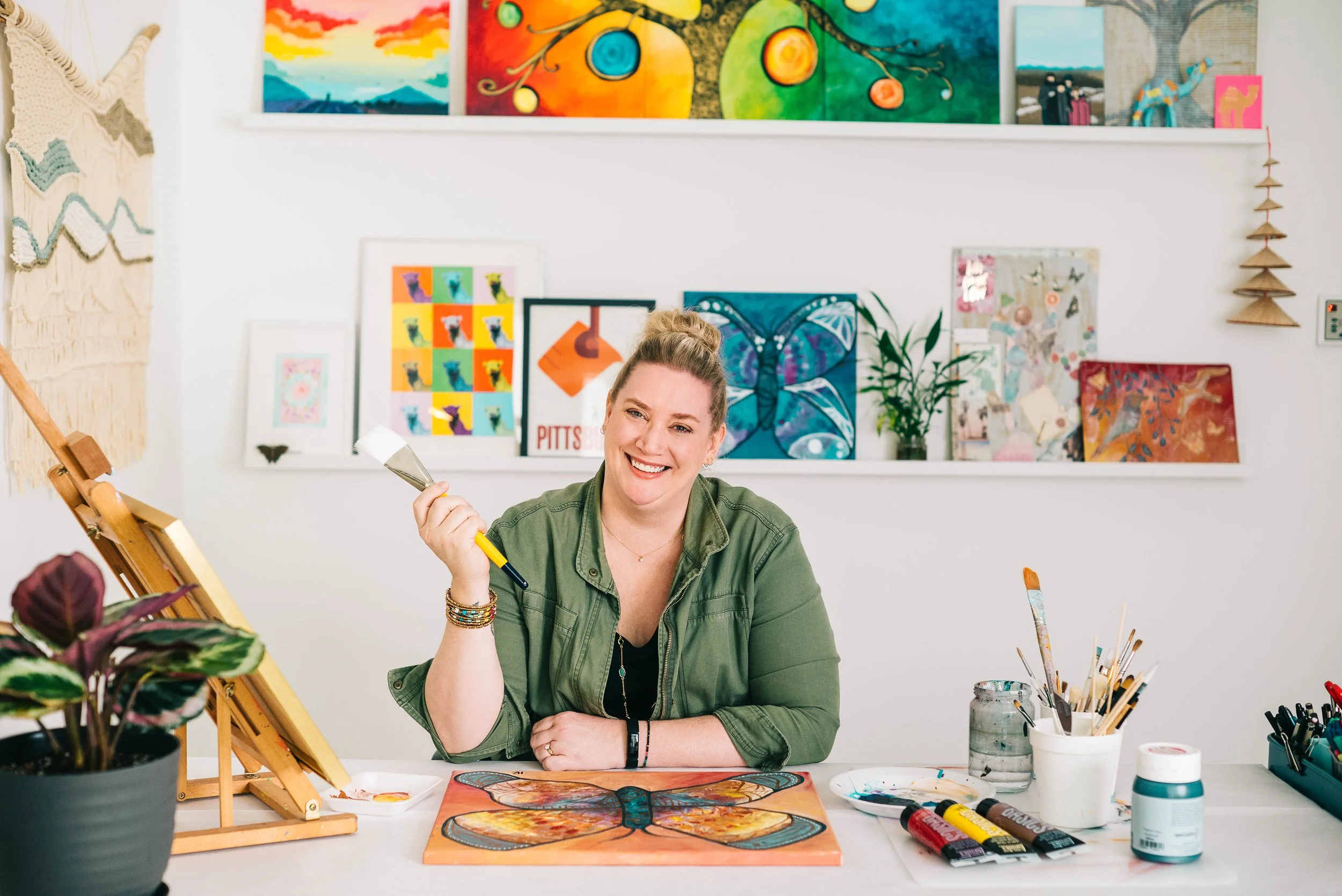 A woman sitting at an art table, holding a palette knife, and smiling at her butterfly painting. The background features shelves with colorful artworks and decorative items.