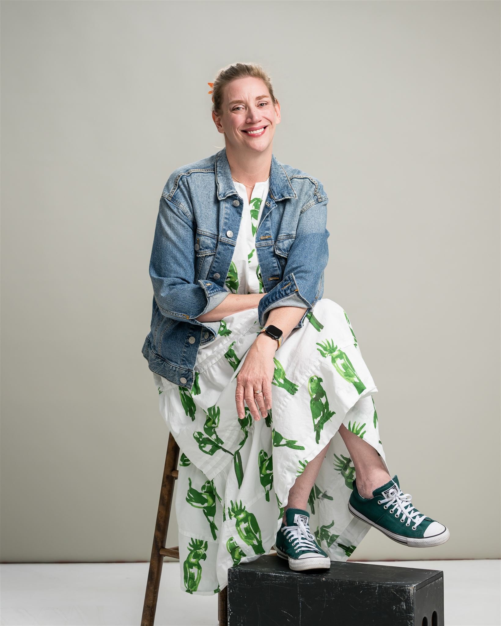 Alt text: Heather Zuchowski seated on a wooden stool with one foot on a black box, smiling warmly in a denim jacket, white green-patterned dress, and green sneakers against a neutral studio background.