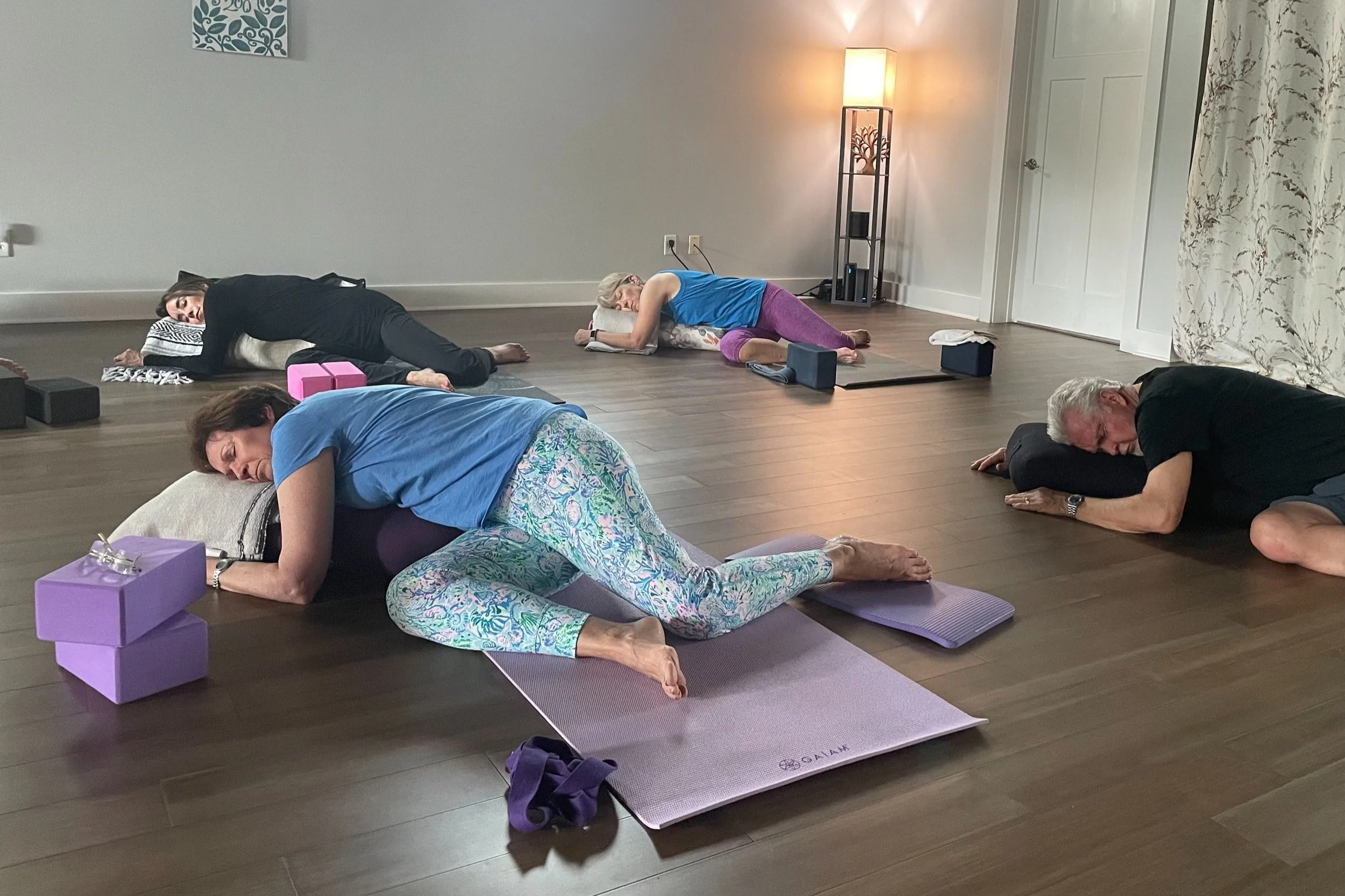 A restorative yoga class, students lying on their sides resting on bolsters, in a spacious and serene yoga studio