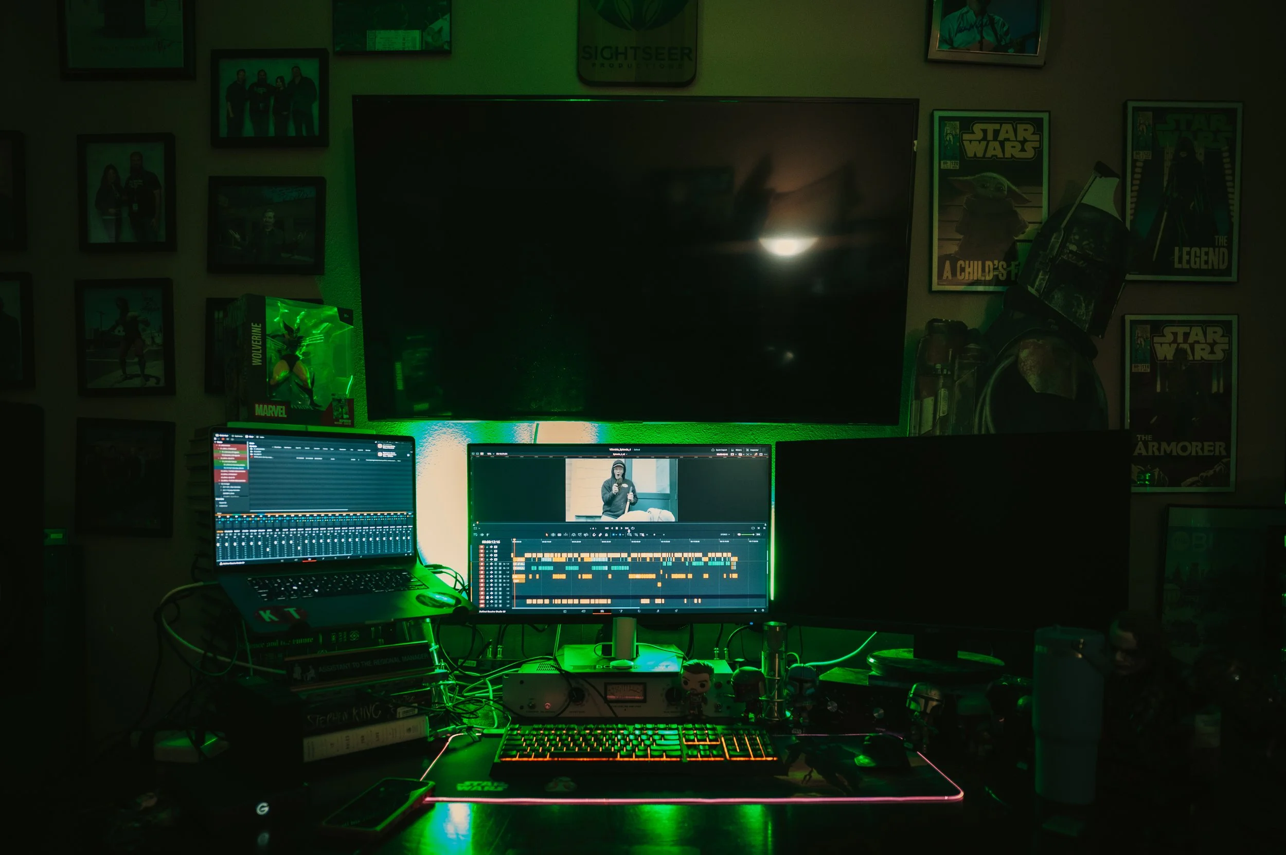 A home music production setup with a keyboard, a laptop, and a monitor displaying a music editing software. Green LED lighting highlights the workspace, which includes books, Funko Pop figures, and various electronic equipment. The background feature