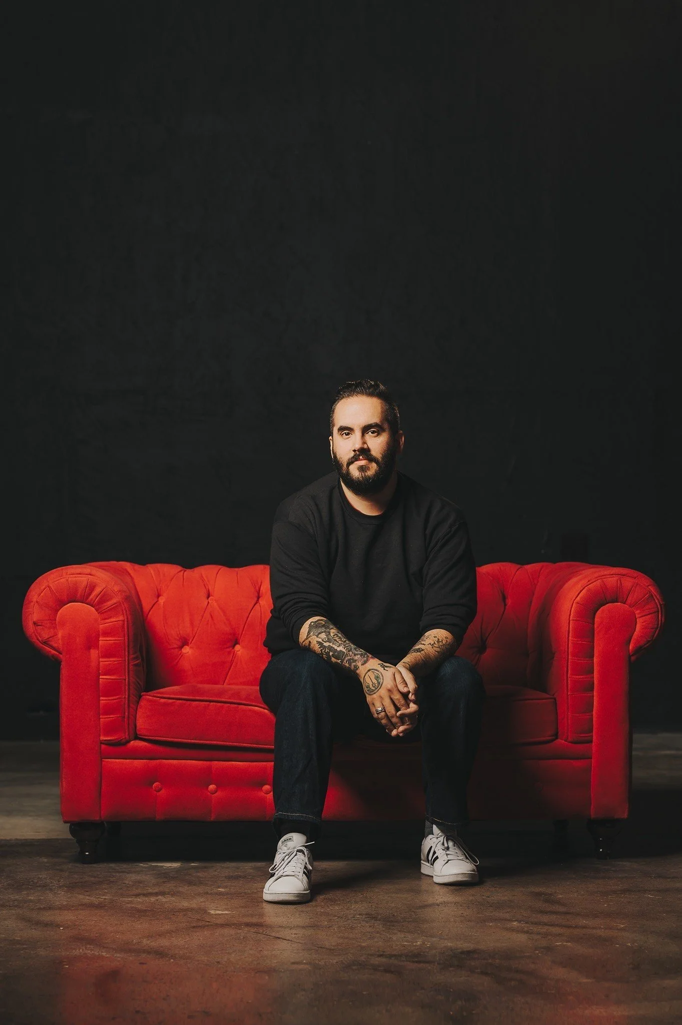 A man with tattoos on his arms sitting on a red sofa against a black background.