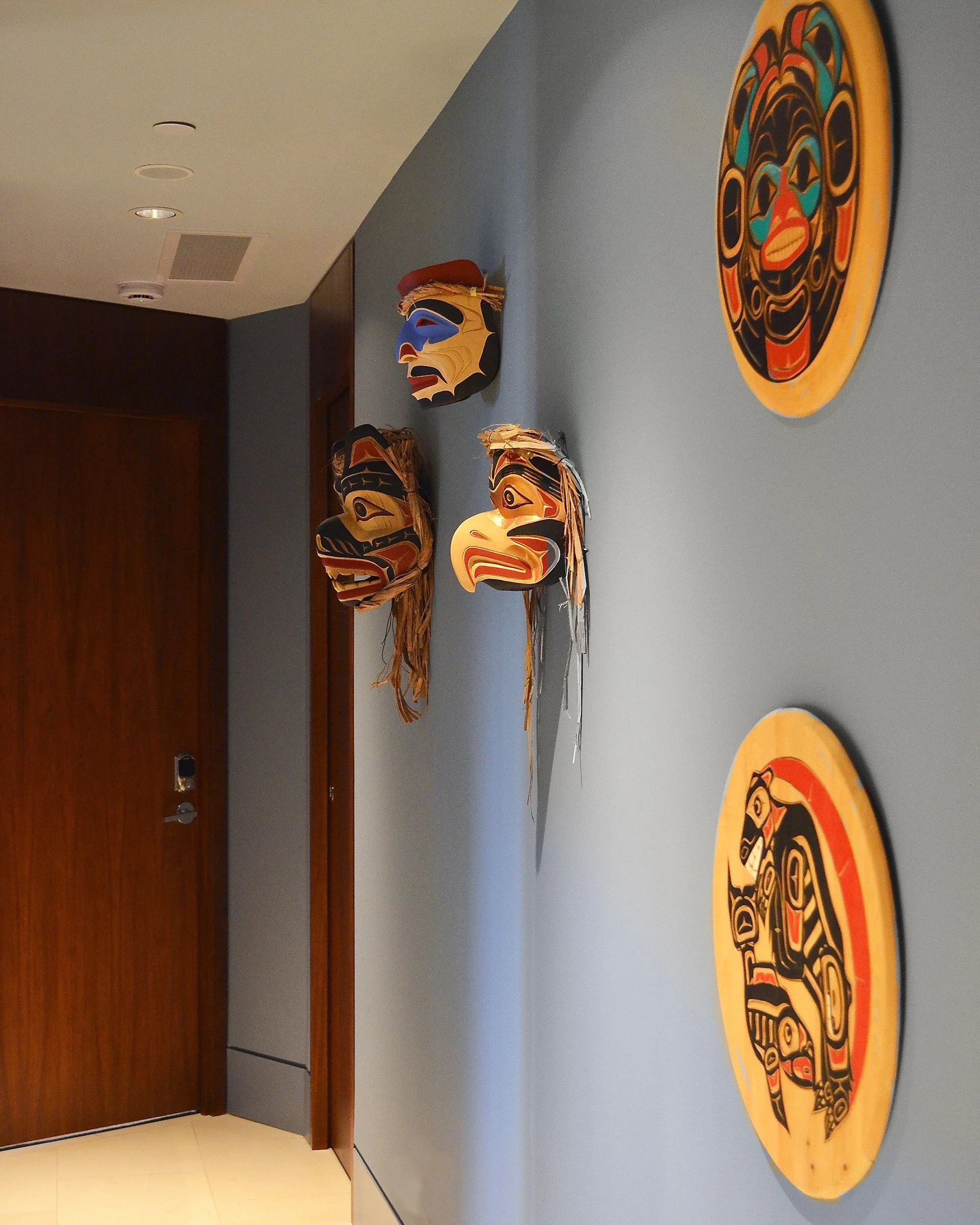 A sophisticated blue-grey hallway provides a quiet backdrop for a collection of Northwest Coast Indigenous masks and carvings, allowing the warmth and vivid colours of the artwork to take centre stage.
