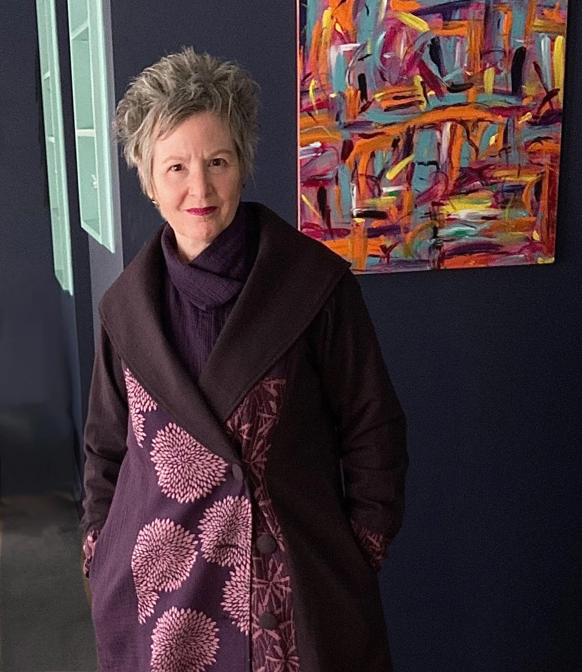 woman in purple coat with flower pattern against dark wall with colourful abstract painting