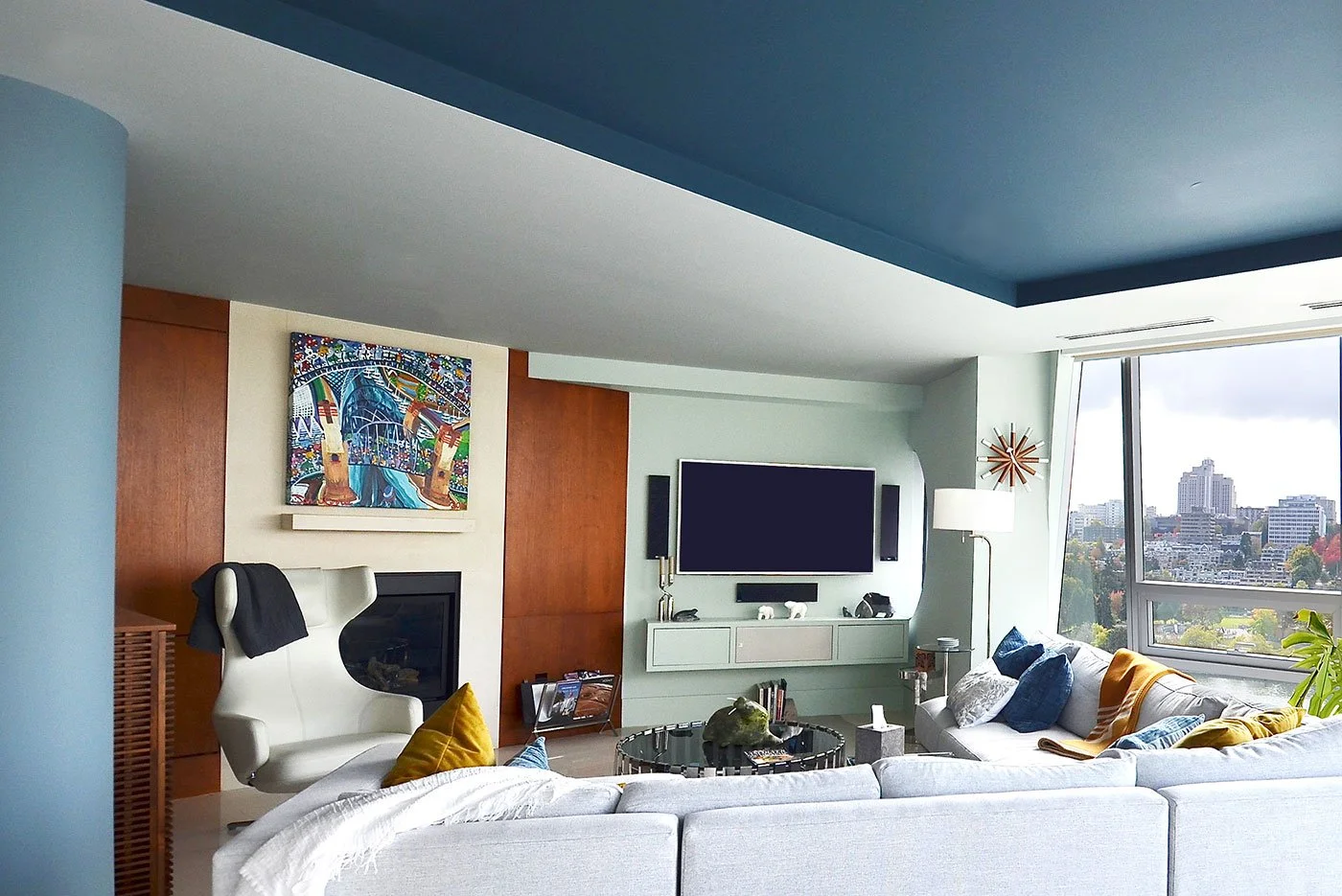 A soft green/blue palette creates a calm, cohesive backdrop for warm wood, white furnishings, and accents of deep blue while connecting the interior to the expansive city view.
