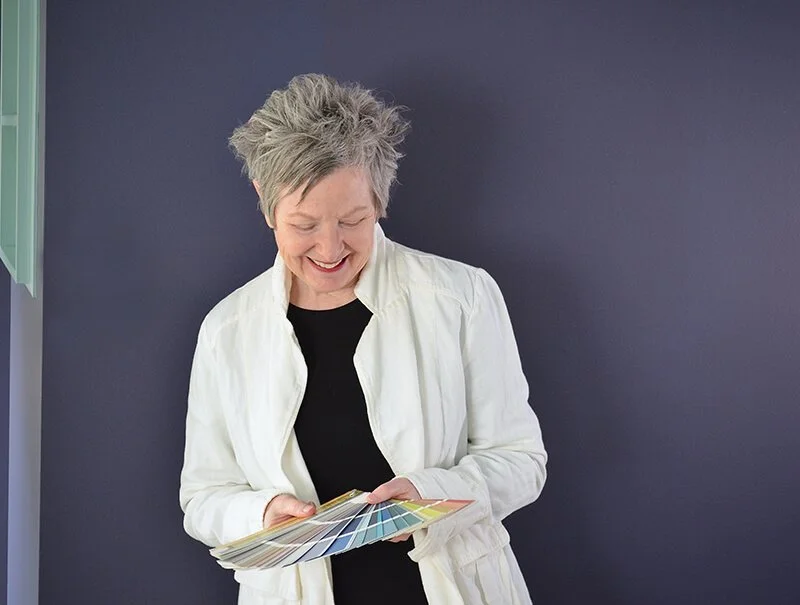 Woman looking down at a paint colour fandeck which she holds open wearing white jacket with purple wall in background