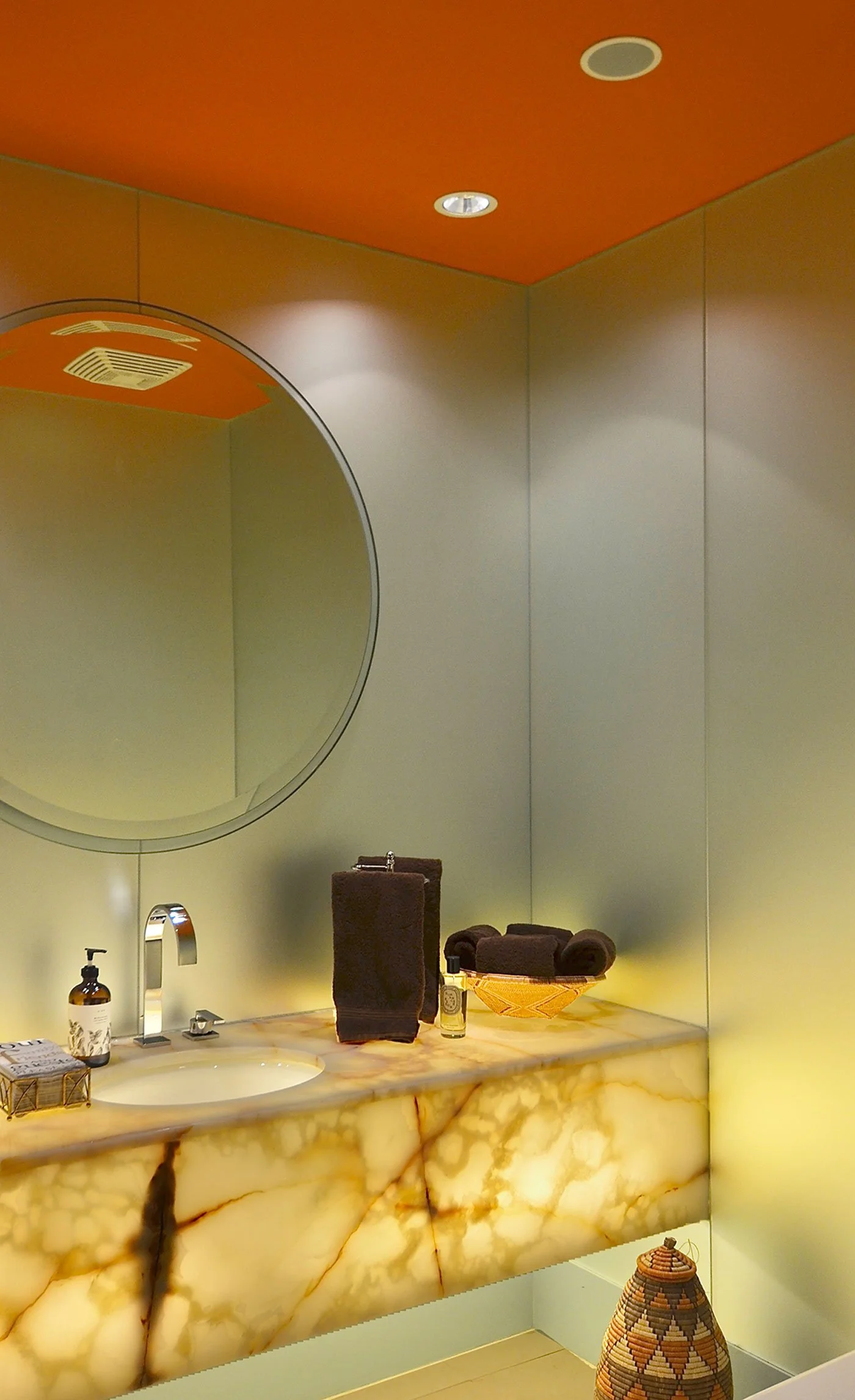 A saturated orange ceiling transforms this intimate powder room into a jewel-like space, creating a warm counterpoint to the softly luminous walls and dramatic veining of the stone vanity. The bold colour brings energy and depth to the compact room, 