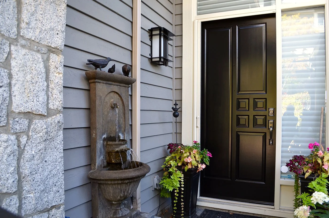 Warm neutrals, natural stone, and a deep charcoal entry door create a sophisticated and welcoming entrance, with the restrained palette allowing the stonework, fountain, and lush planting to add character.