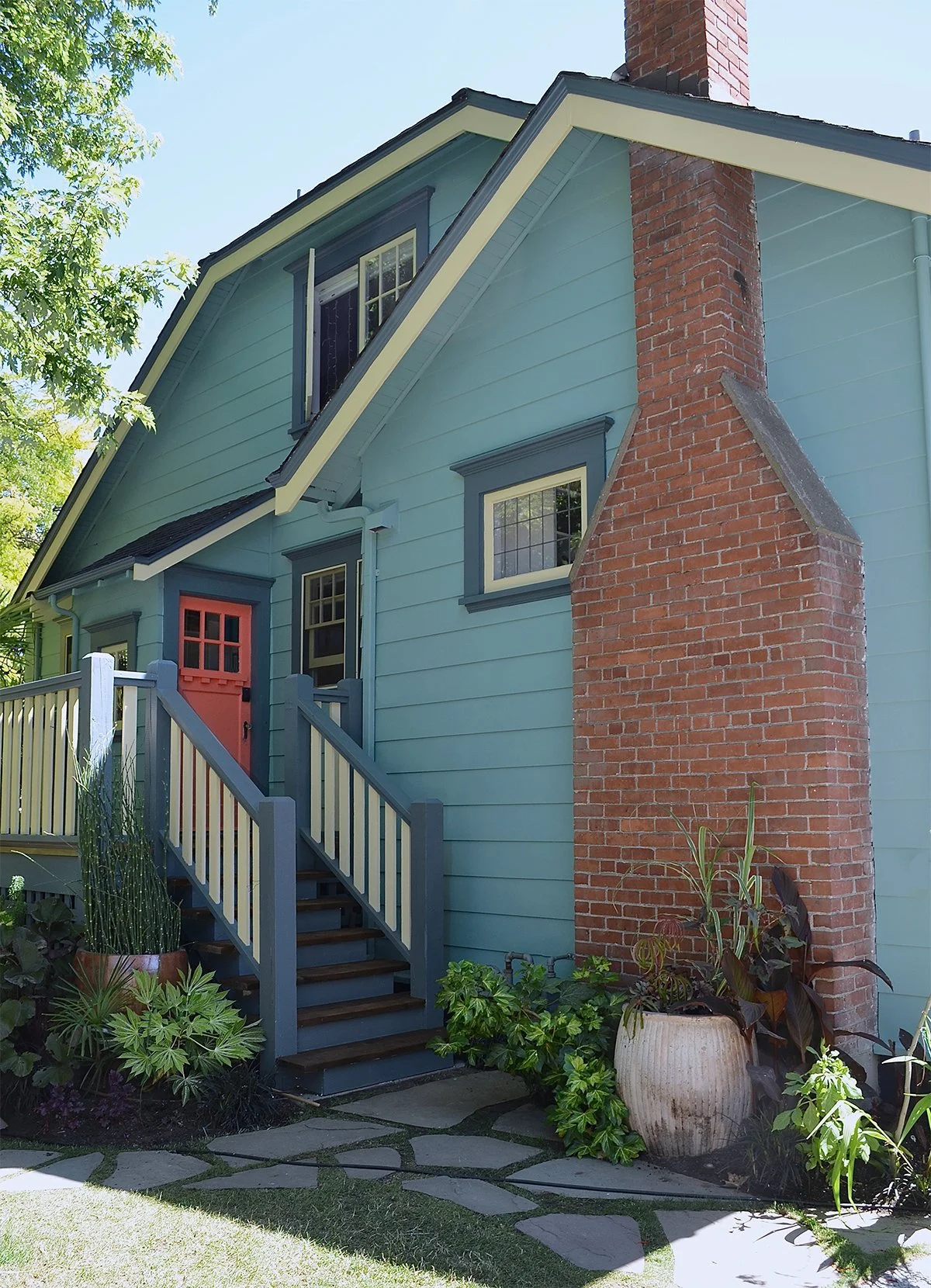 Side entrance of house with green siding, coral door , dark blue trim and yellow windows with brick chimney