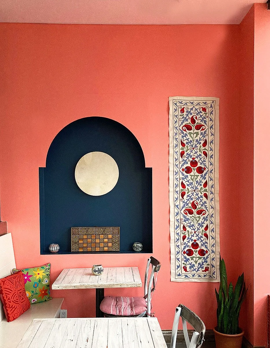 A coral coloured wall with a dark blue arched inset with wall hanging and various objects with tables and chairs in foreground