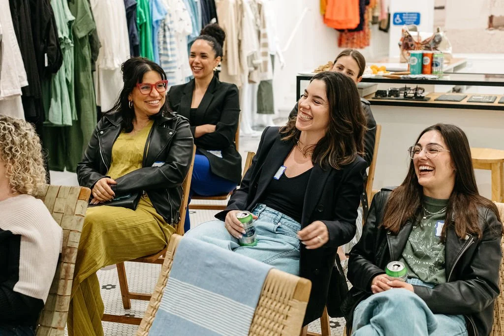 Group of women sitting and laughing together in a clothing store, holding drinks, with racks of clothes in the background.