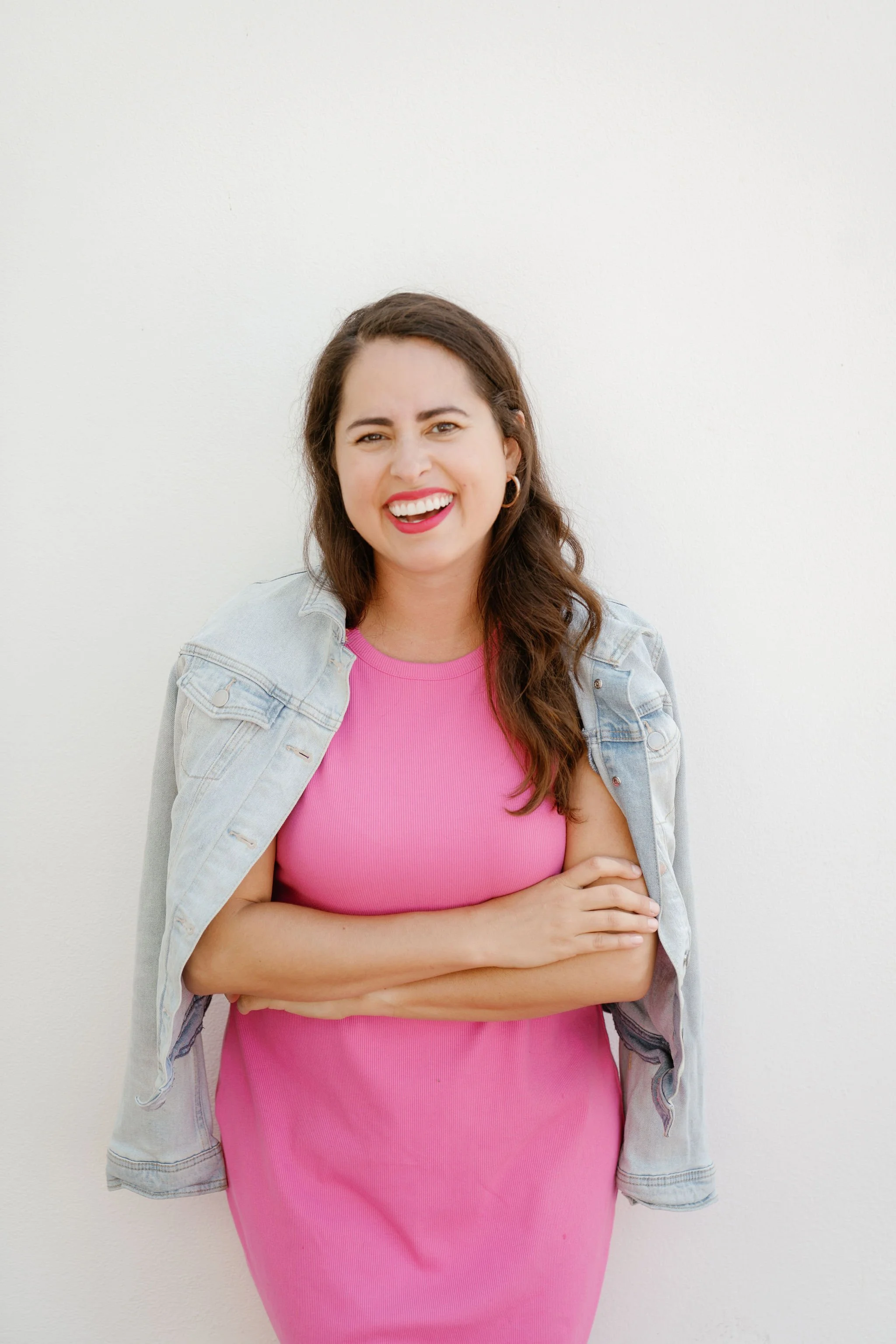 Woman in pink dress and denim jacket smiling against a white wall