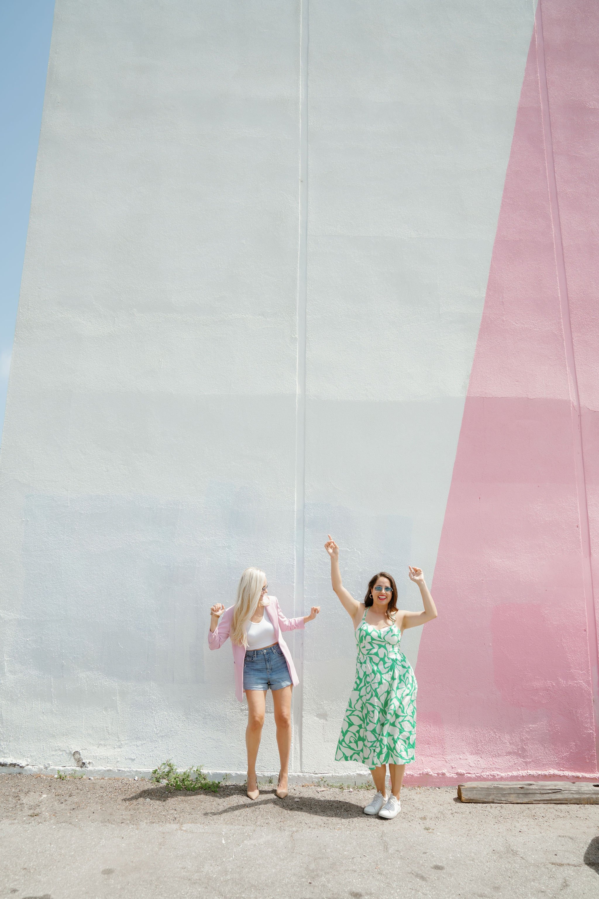 Two women standing in front of a large, colorful mural wall with pastel pink and white sections. One woman wears a pink blazer, white top, denim shorts, and heels, and the other wears a green and white patterned dress with white sneakers. Both women are smiling, with the woman in the dress raising her arms.