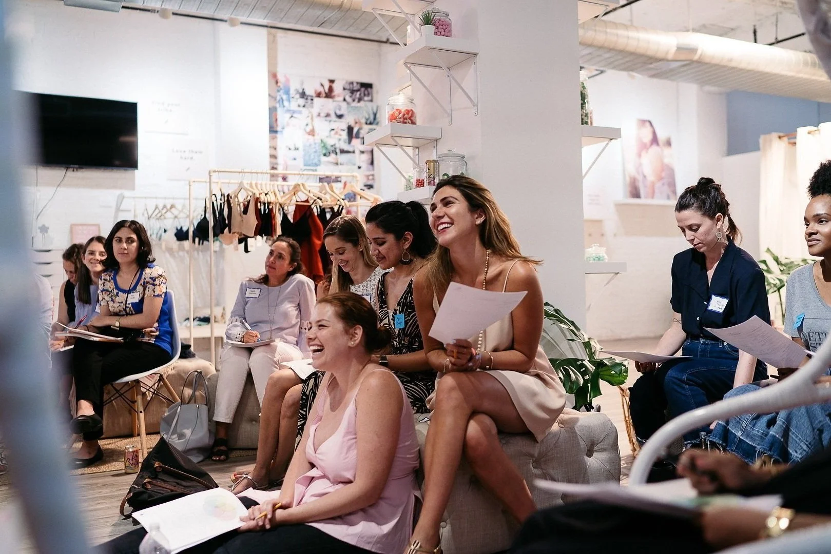 Group of women attending a workshop or seminar, some are smiling and taking notes, in a bright modern space with clothing racks and artwork on the walls.