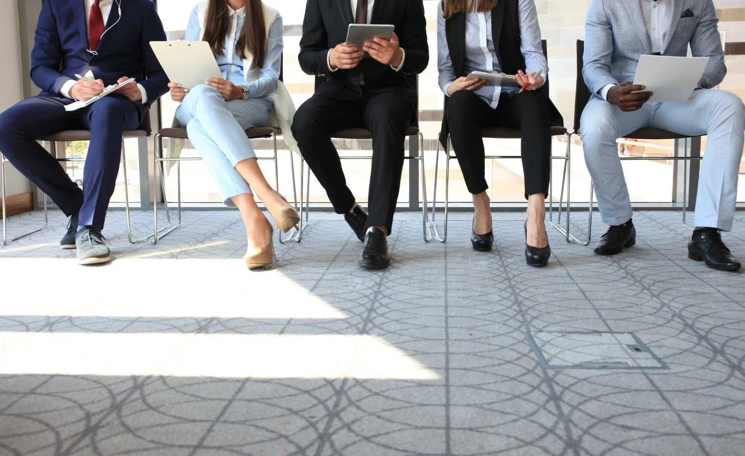 Photo of five people in professional attire sitting on chairs in a row in an office setting, visible from the chest down. They are holding clipboards and filling out forms.
