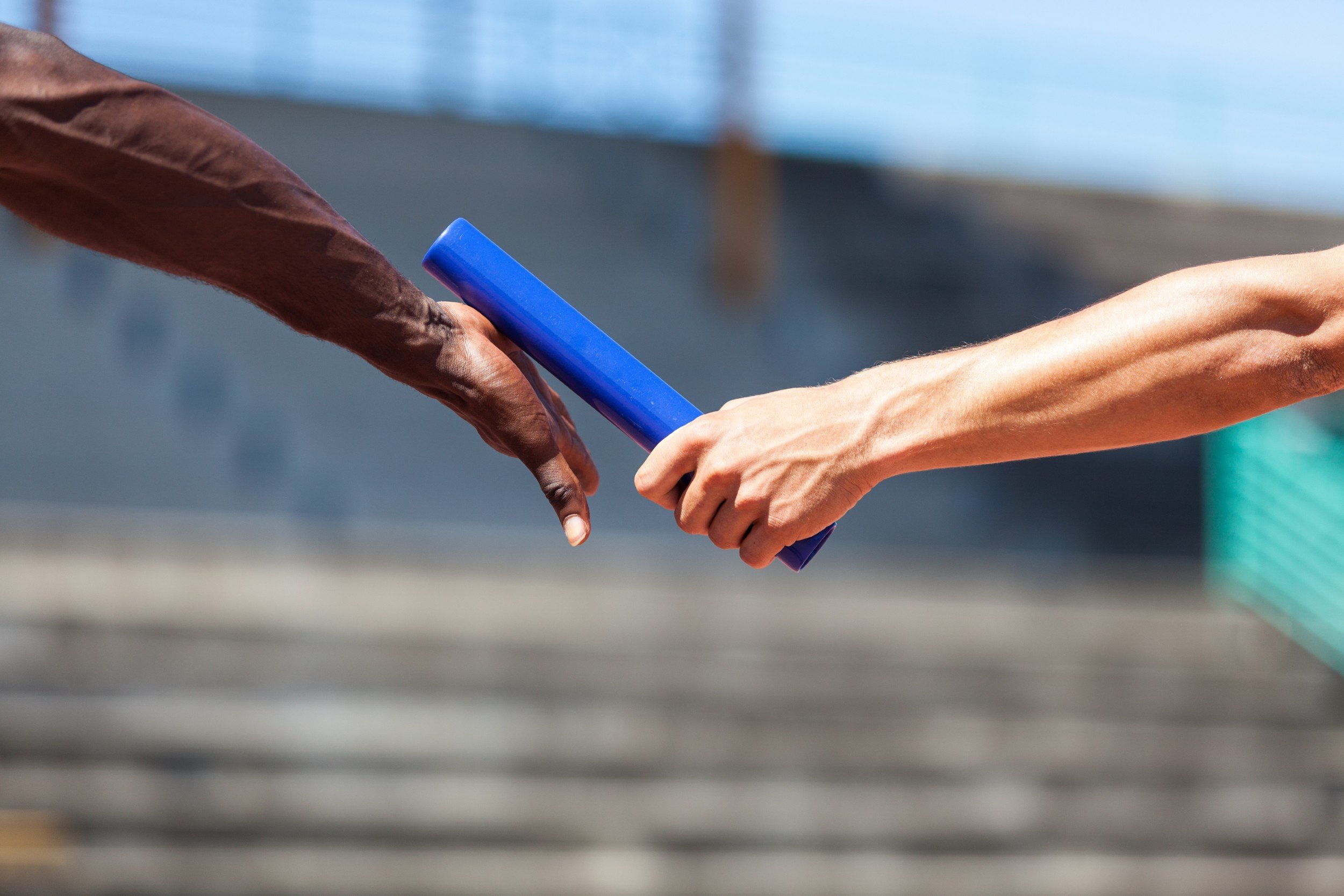 In the foreground are the hands and forearms of two people who appear to be runners in a relay race. On the right, a white person's hand is extended with a blue baton in hand, passing it to the outsteched palm of a Black person's hand.