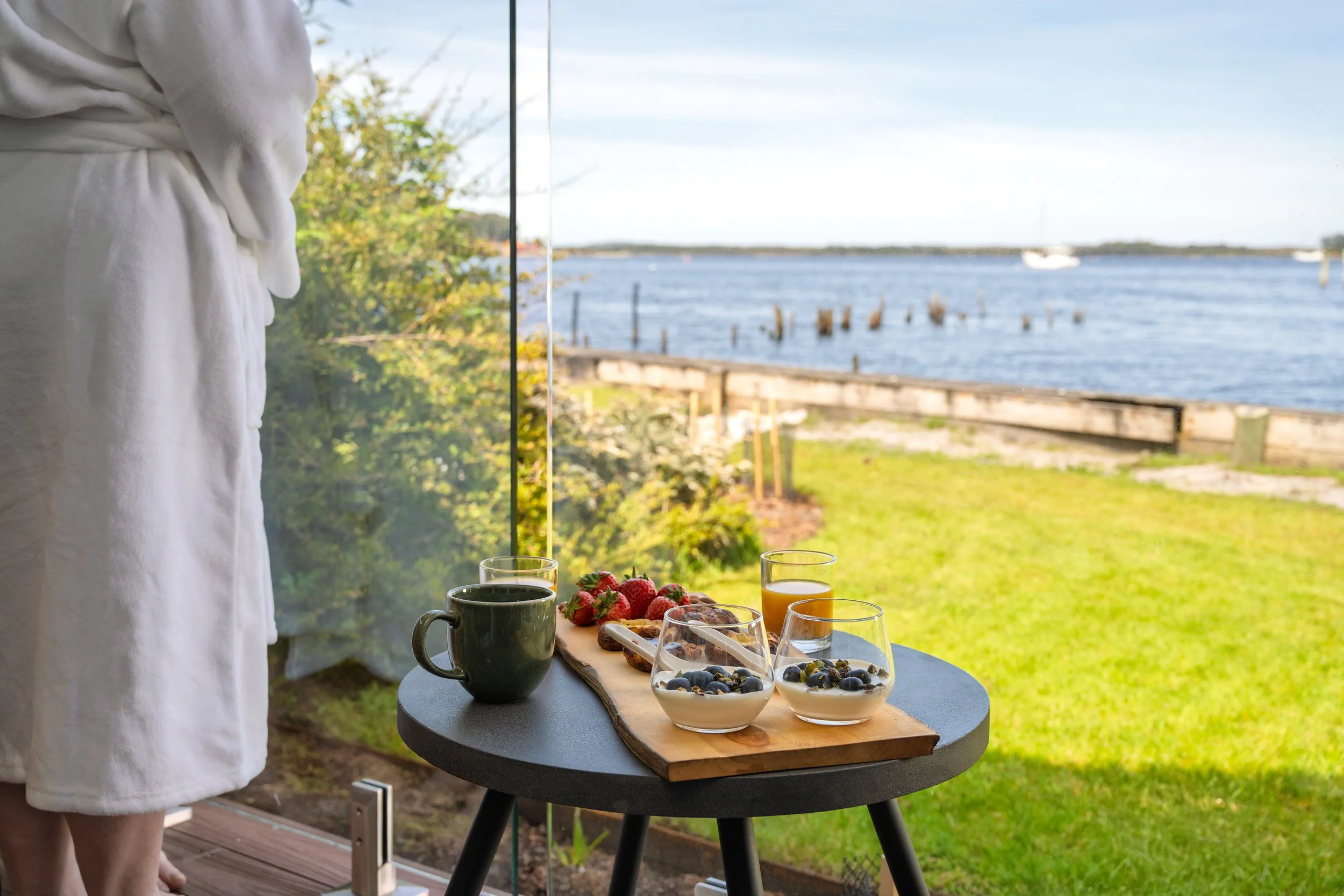 Breakfast table with fruit, coffee, and juice near a large window overlooking a grassy yard and a body of water with boats in the background.