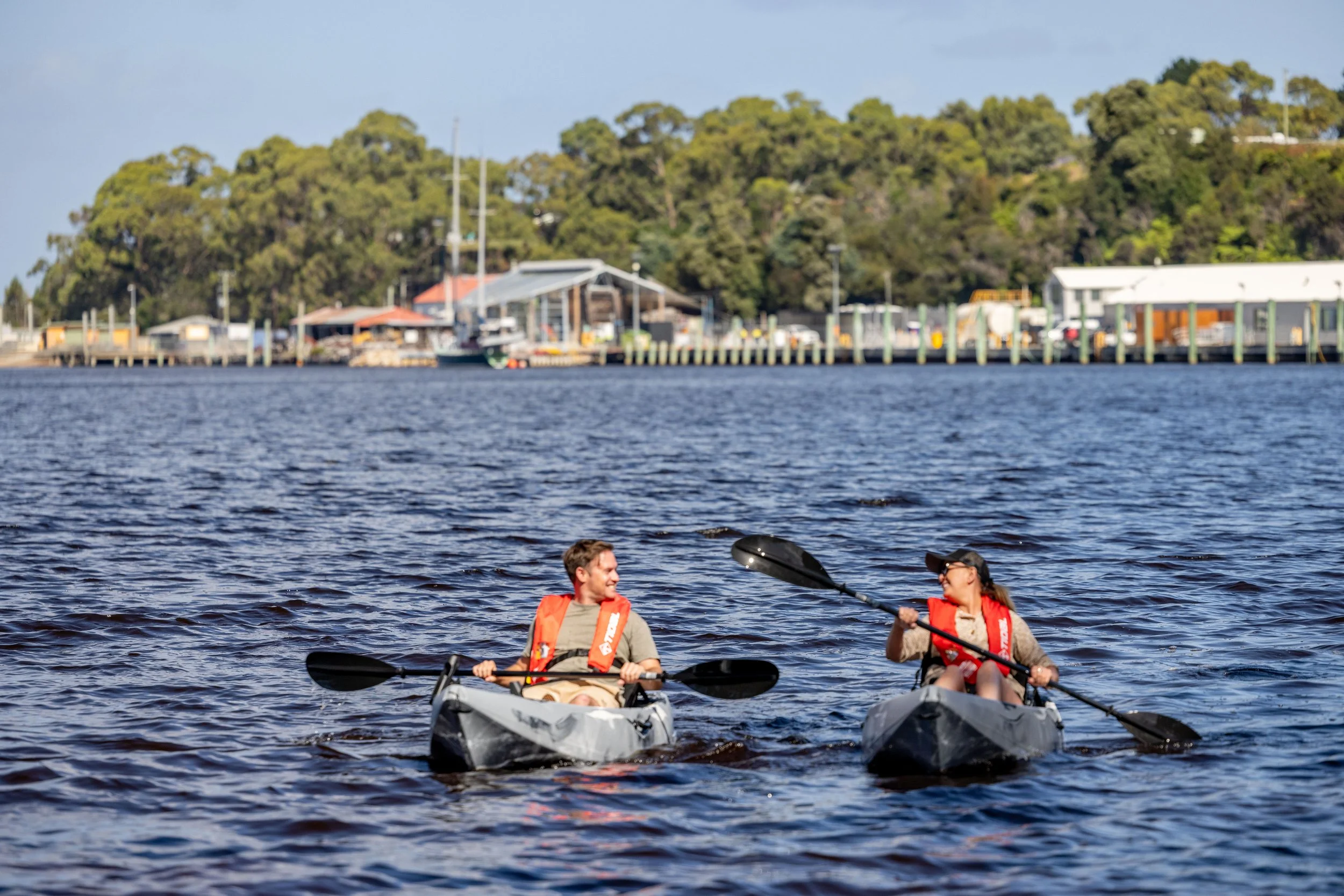 Two people kayaking on Macquarie Harbour with trees and buildings in the background.