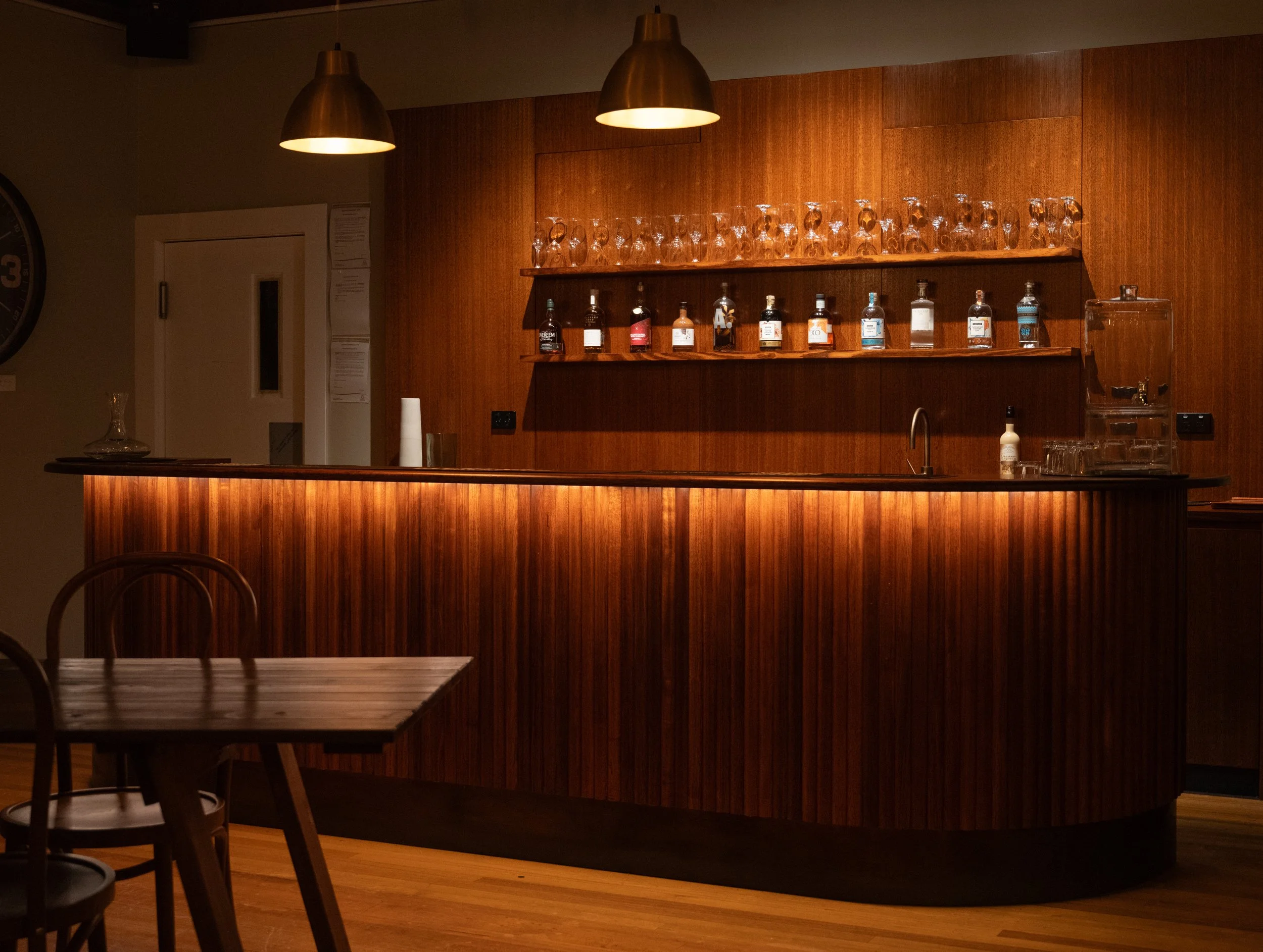 A Tasmanian Oak bar counter with bottles of alcohol and wine glasses on shelves behind it, illuminated by warm lighting from above, in The Mill at Risby Cove