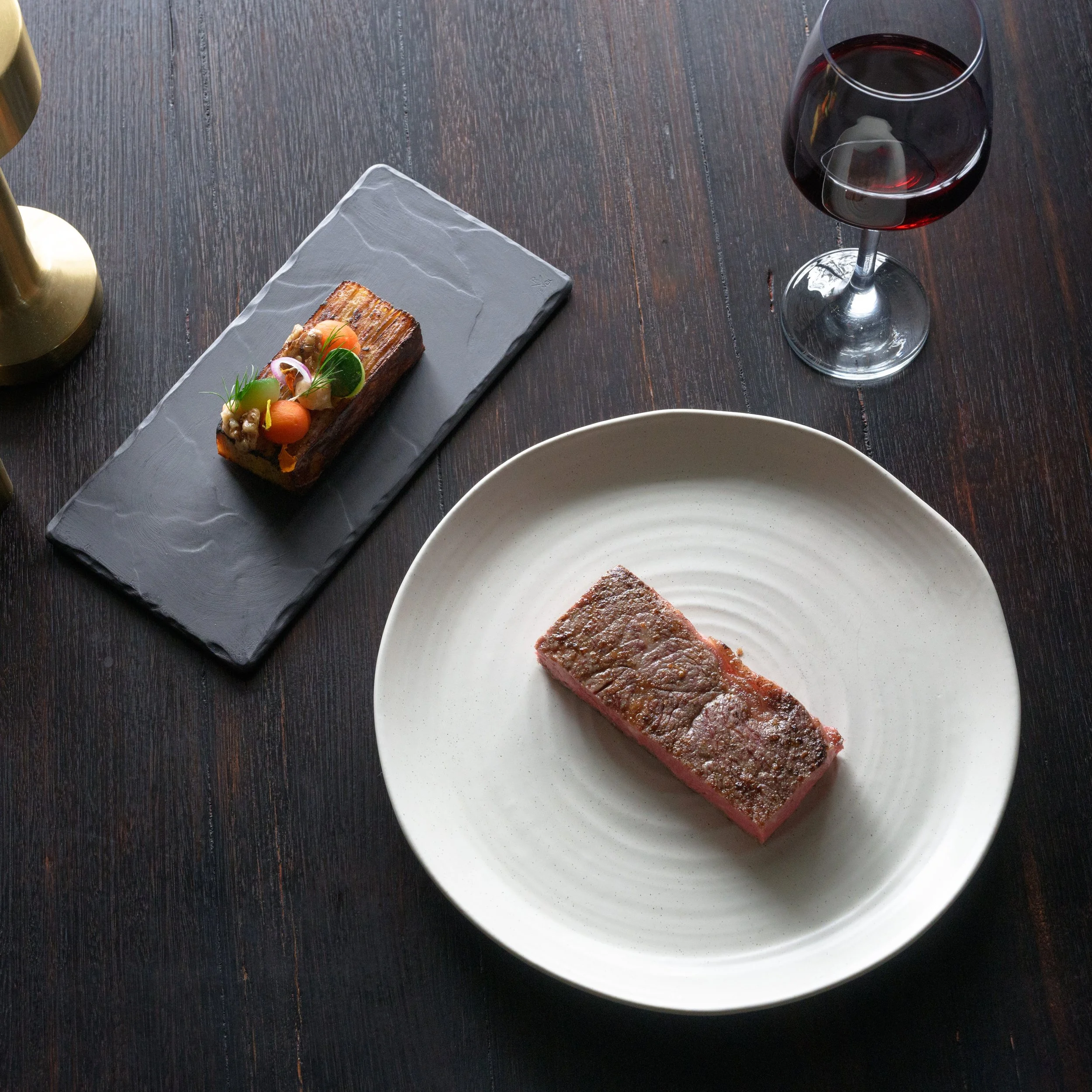 A plate with a Robbins Island MB9+ Wagyu Scotch Fillet, with a side of mille feuille topped with microgreens and vegetables, and a glass of red wine.