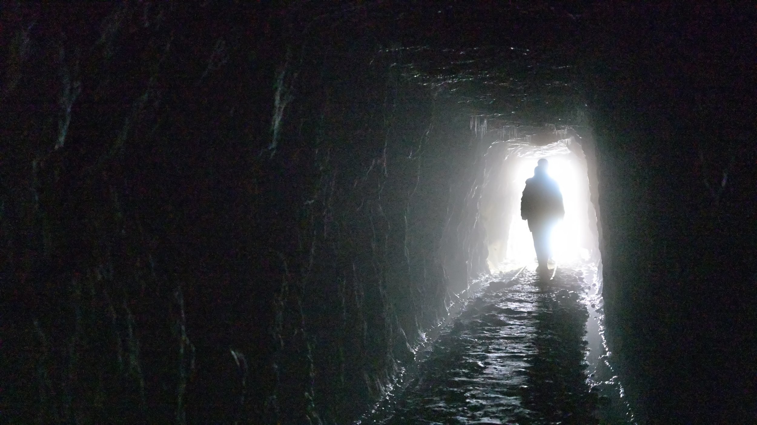 Person walking through a dark, rocky mine shaft with light shining at the end.