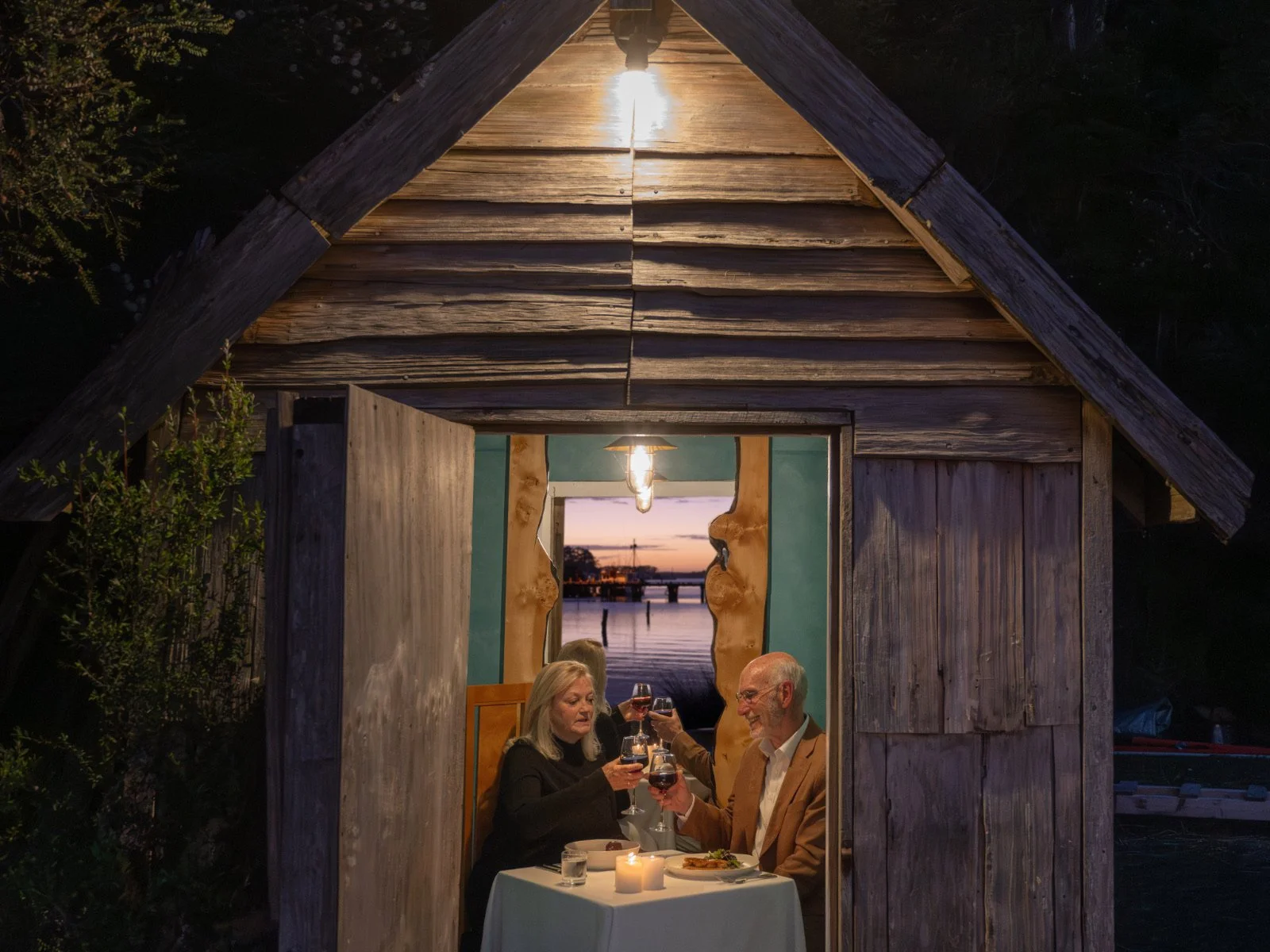 A couple dining inside an old Piner's Hut on the edge of Macquarie Harbour at sunset, with a candle and glasses of red wine on the table.