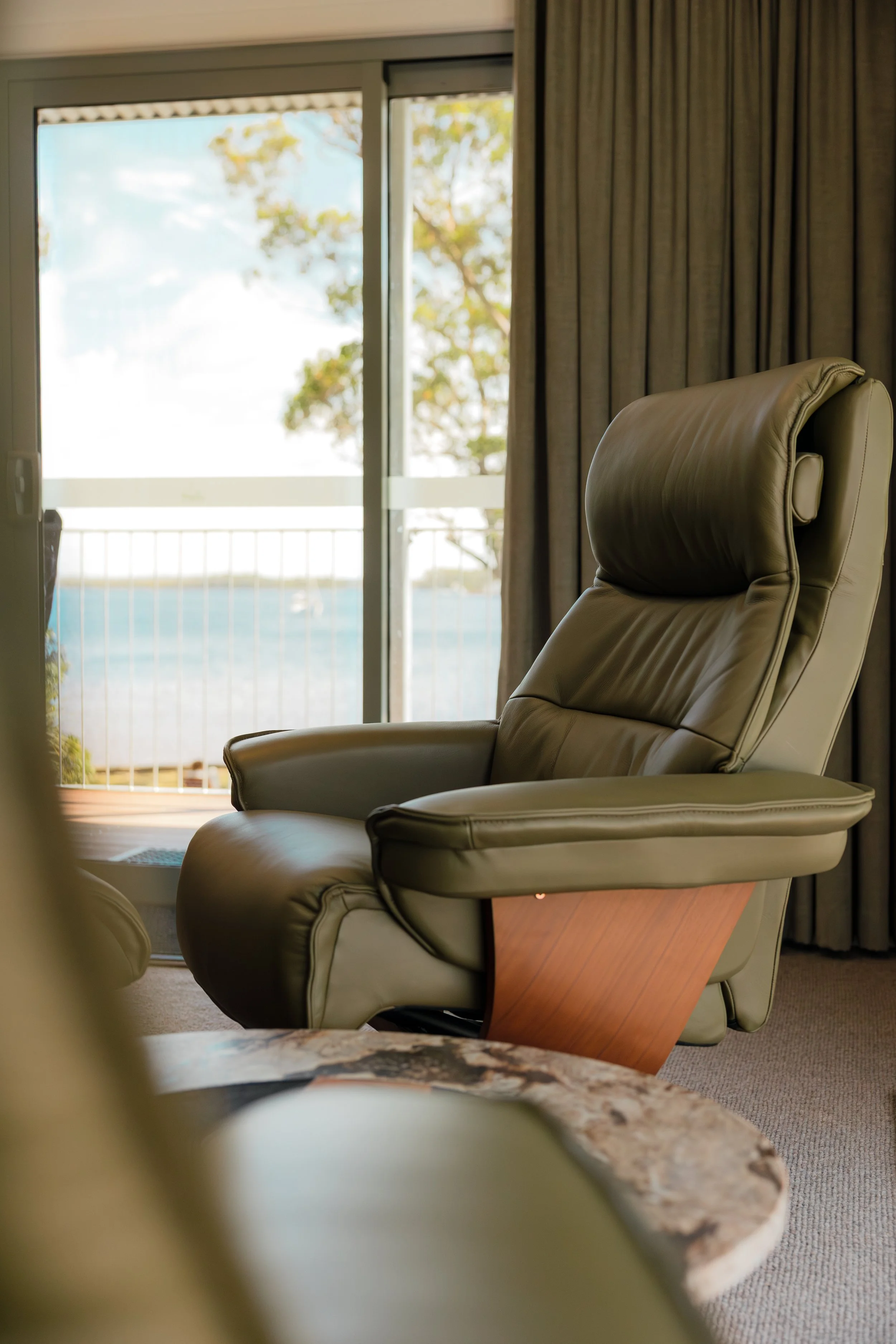 A green leather recliner chair in a living room with a large window and curtains, overlooking a balcony and a body of water with trees in the background.