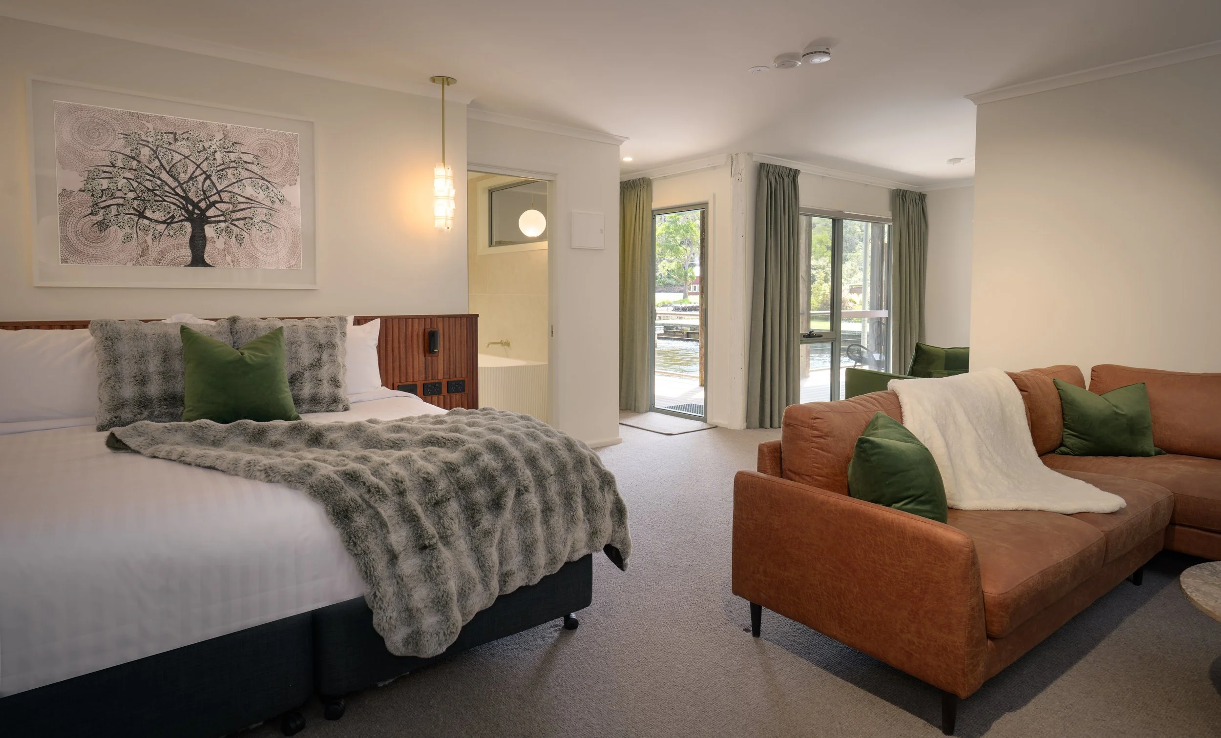Hotel room with a bed, green and gray pillows, a fur throw, and artwork of a tree; living area with a brown couch, green pillows, blanket, and sliding glass doors leading to an outdoor area.