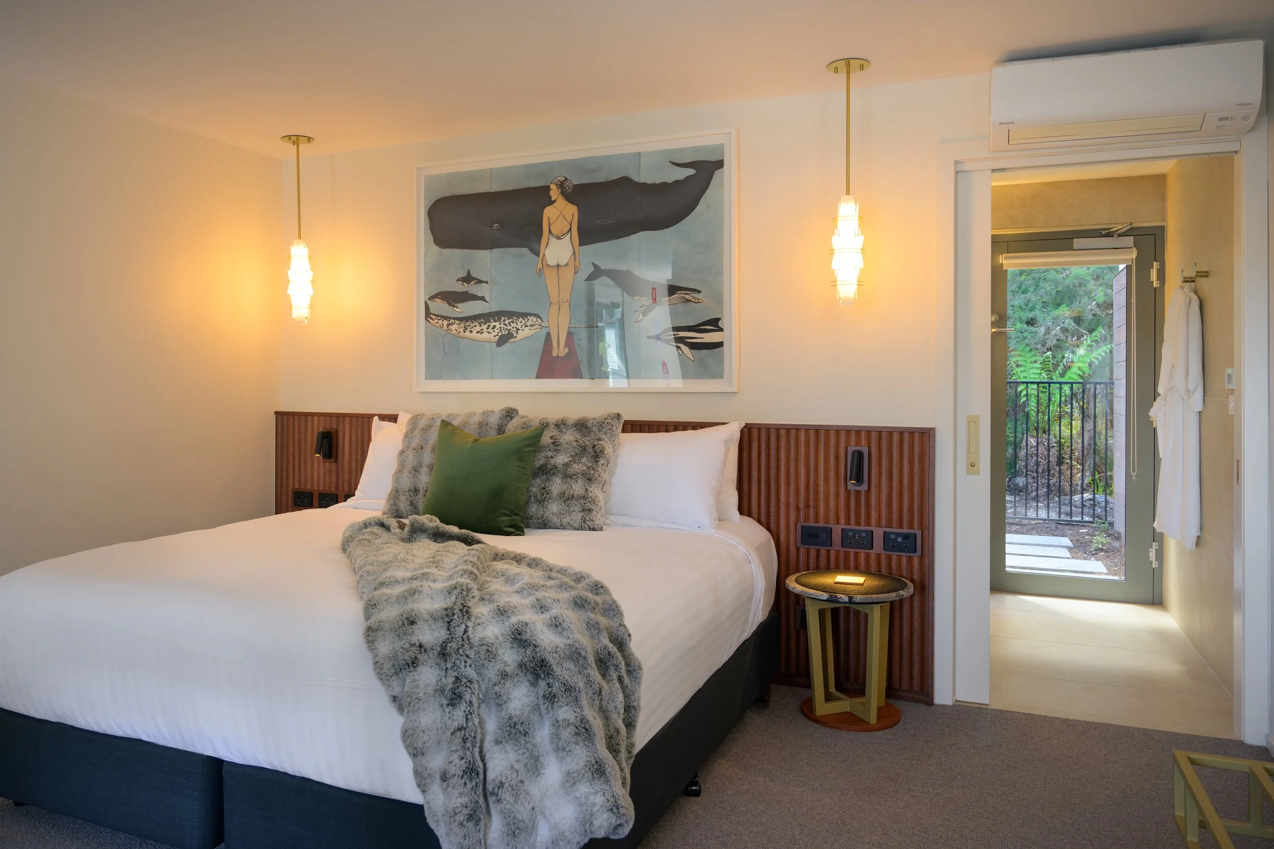 Modern bedroom with a large bed covered in white linens, surrounded by decorative pillows and a faux fur throw. Two hanging pendant lights illuminate the room, and a framed painting of a woman with marine animals hangs above the bed. There is a door leading outside showing greenery, and a wall-mounted air conditioning unit.
