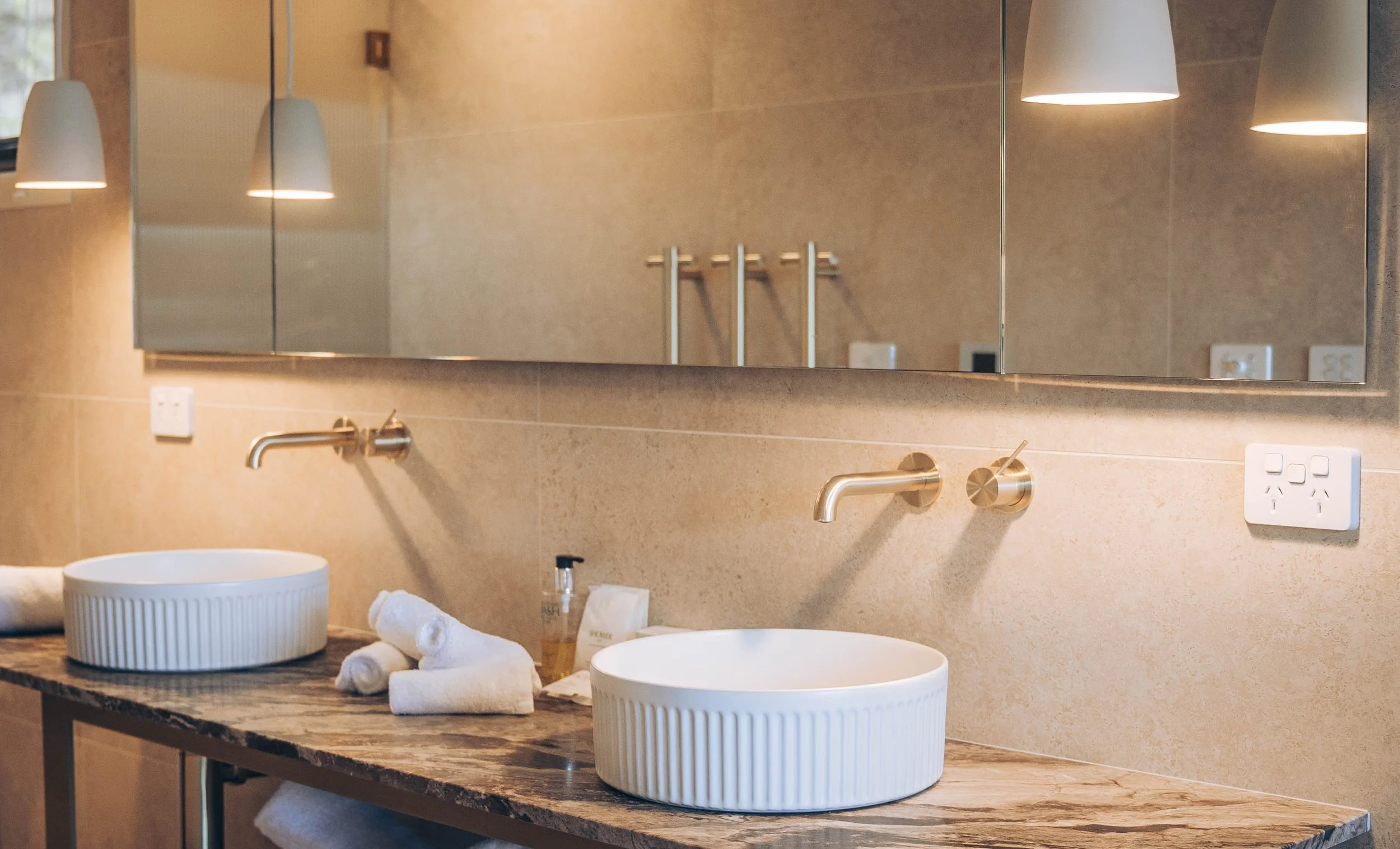 Modern bathroom interior with a marble countertop, dual round sinks, wall-mounted faucets, large mirrored wall, pendant lighting, and rolled towels.